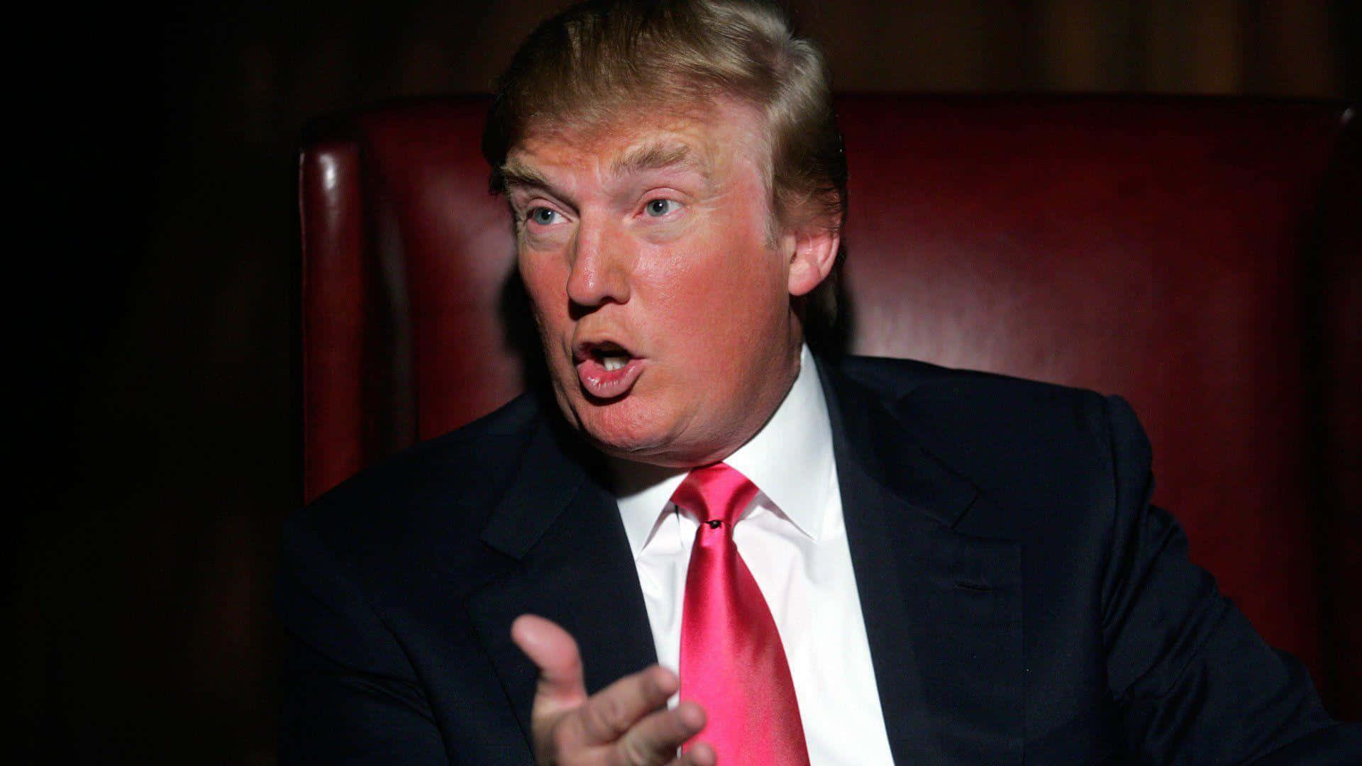 trump 桌布 壁紙 — 一名身穿深色西裝和鮮紅色領帶的男子坐在紅色皮椅上，一邊說話一邊做手勢，表情強烈。