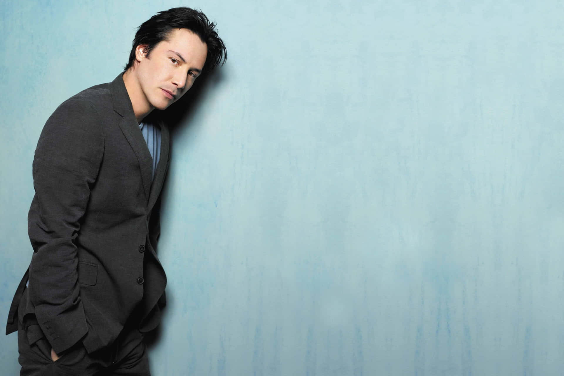 Keanu Reeves 桌布 壁紙 — 一名身穿深色西裝的男子，表情嚴肅地靠在一面有紋理的淺藍色牆上。