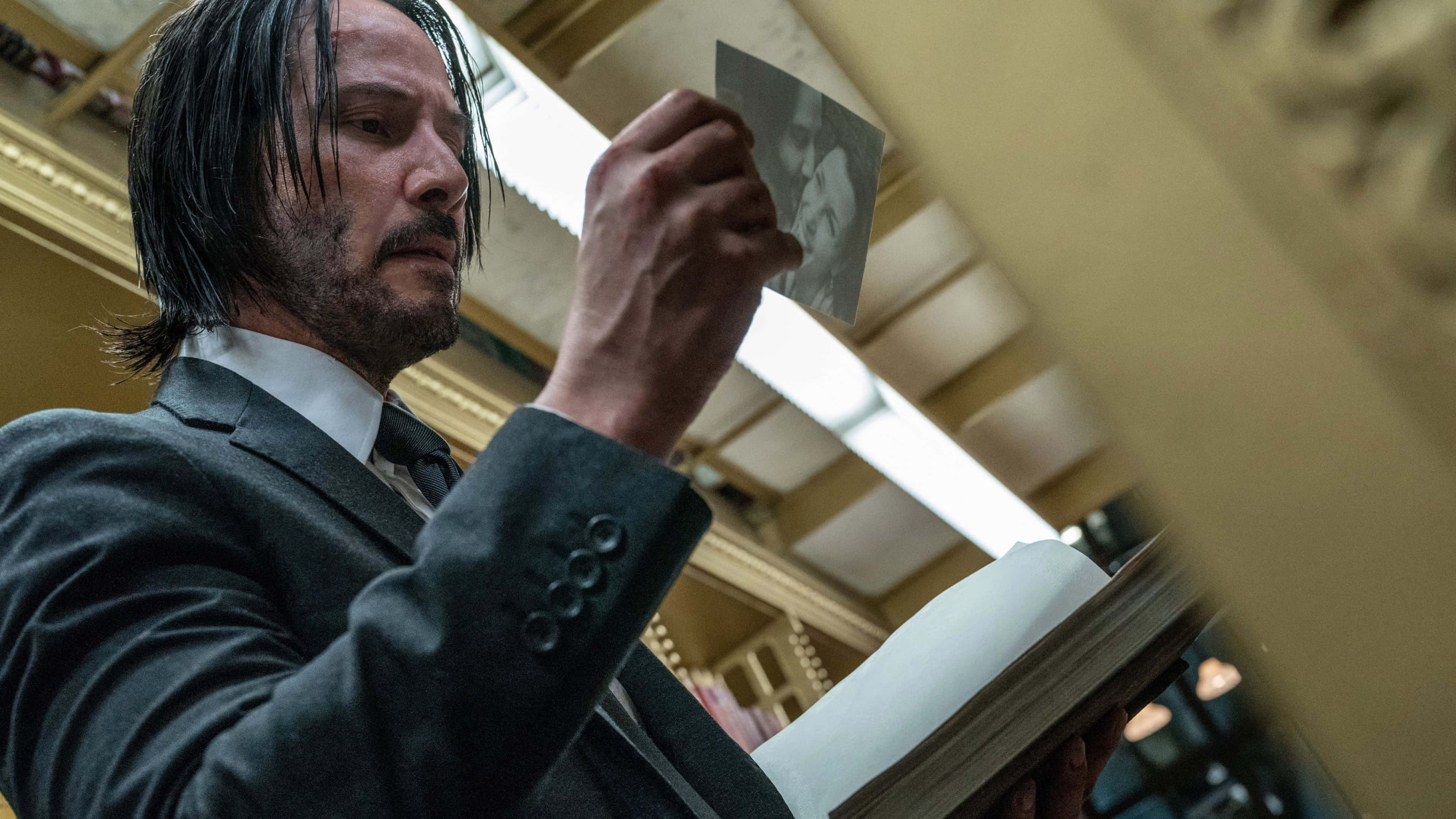 Keanu Reeves 桌布 壁紙 — 一名身穿深色西裝的男子在一個裝飾華麗、燈光溫暖的大房間裡，將一張照片舉在一本打開的書上方。