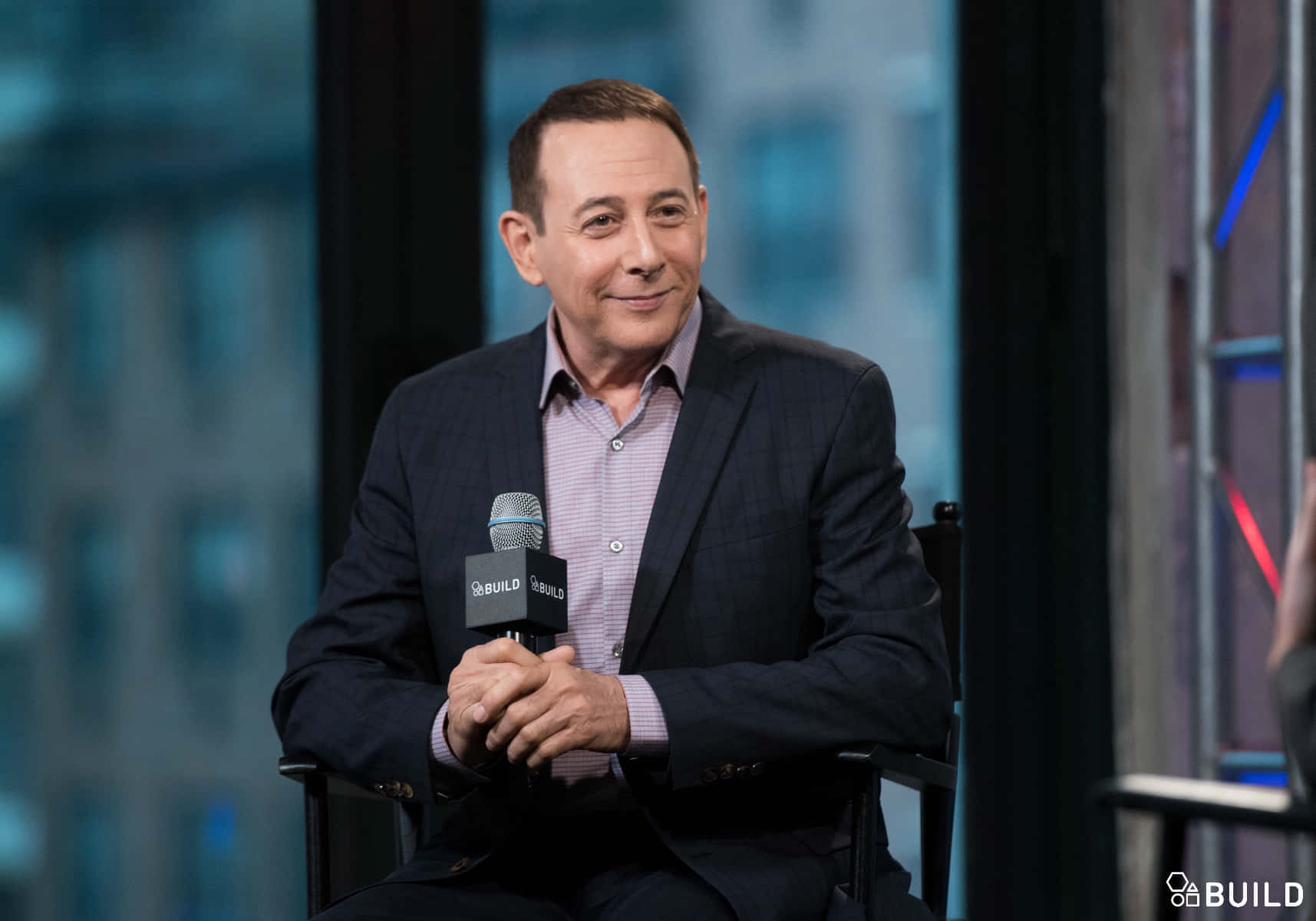Paul Reubens 桌布 壁紙 — 一名身穿深色西裝的男子在現代化錄影棚內接受採訪，手持麥克風微笑，背景是模糊的城市天際線。
