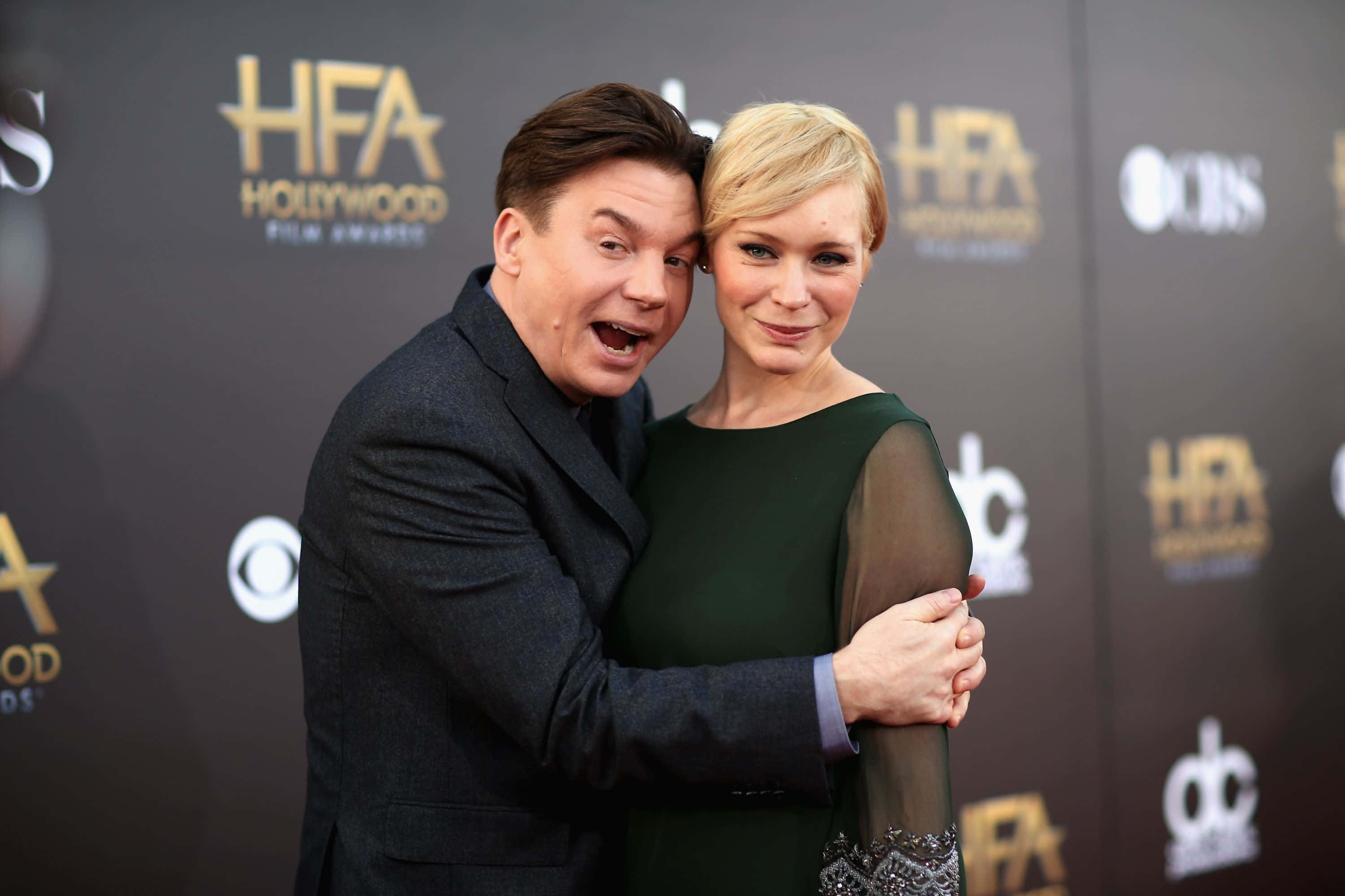Mike Myers 桌布 壁紙 — 一名身穿深色西裝的男子在荷里活電影獎紅地毯活動上擁抱一名身穿綠色連衣裙的女子。