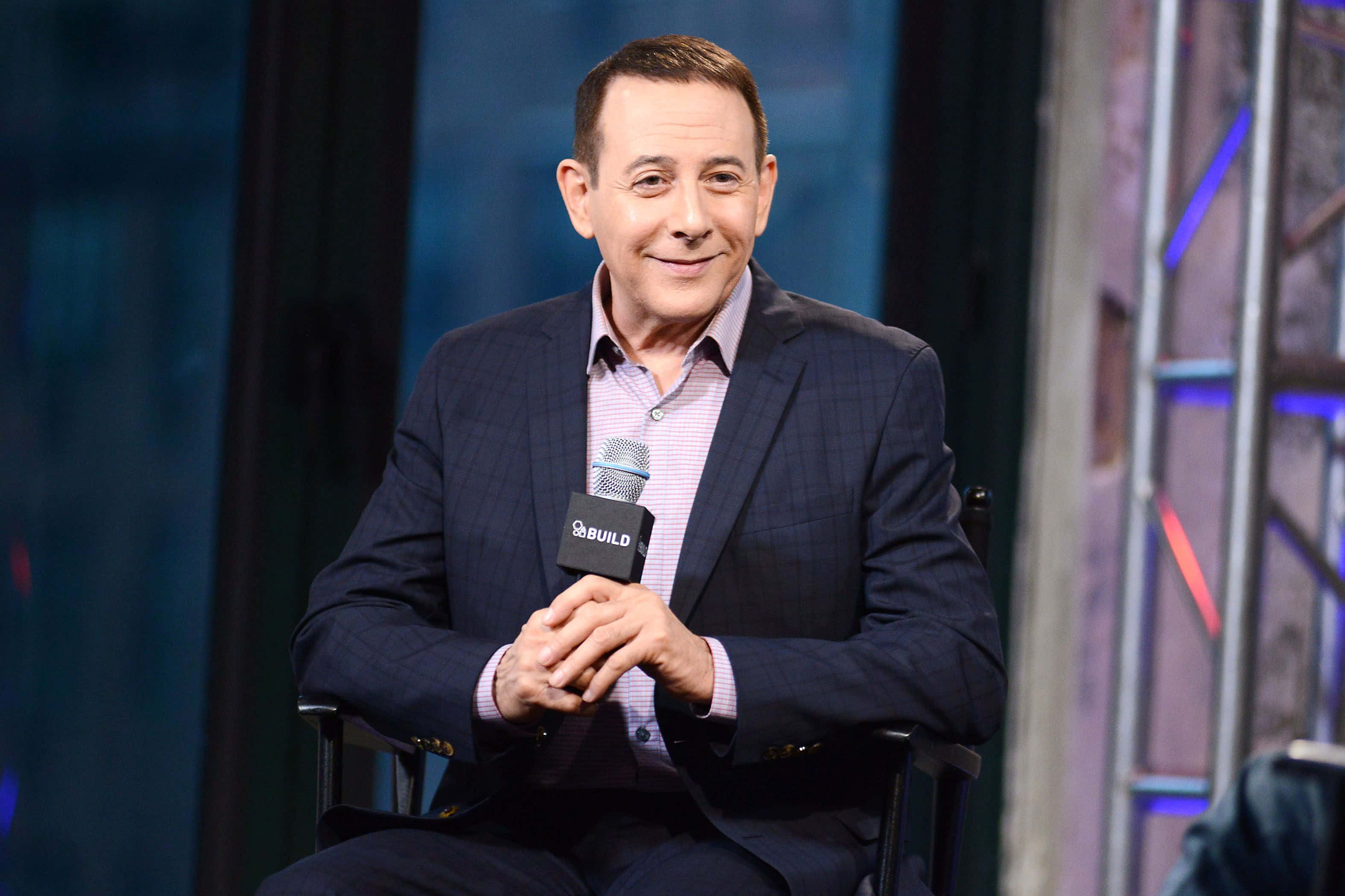 Paul Reubens 桌布 壁紙 — 一名身穿深色西裝的男子在藍色和紫色燈光的攝影棚中微笑著手持麥克風。
