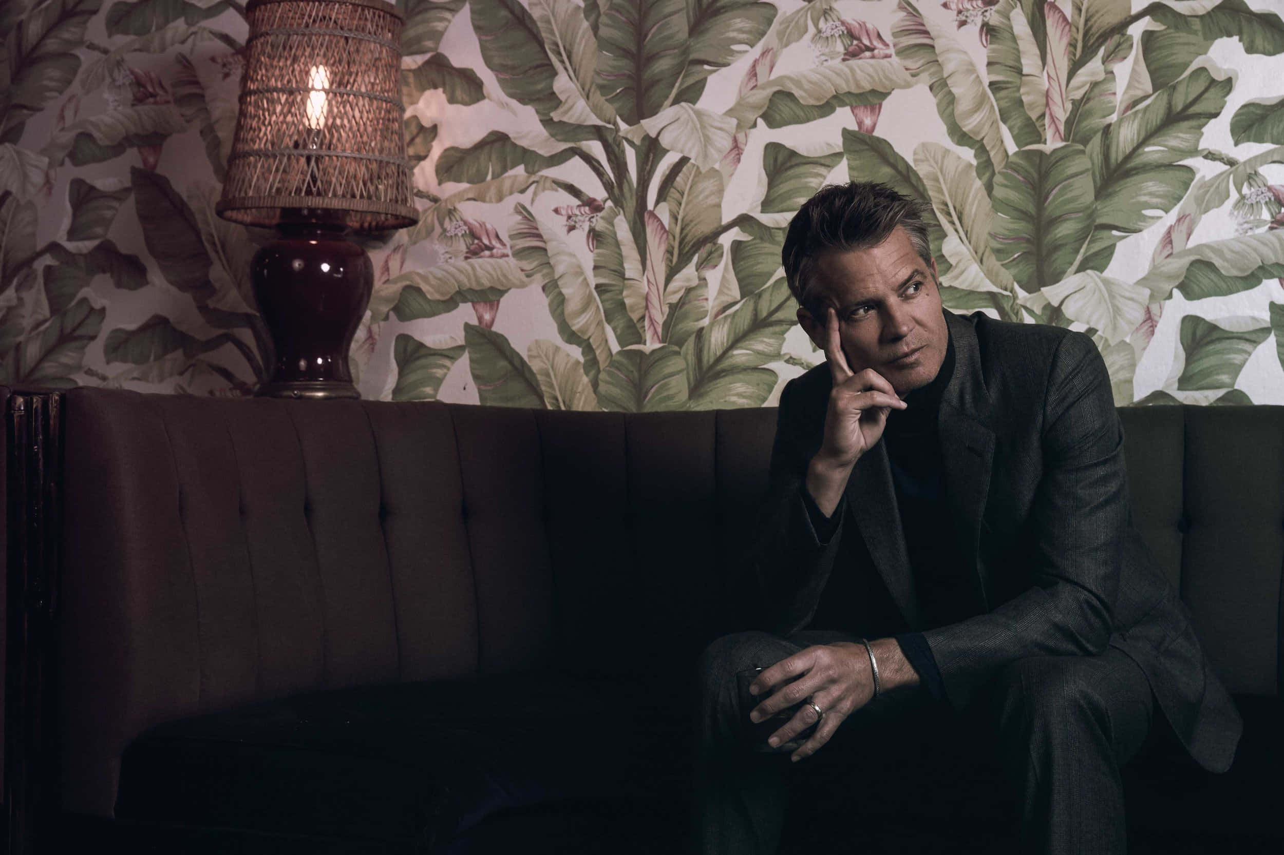 timothy olyphant 桌布 壁紙 — 一名身穿深色西裝的男子坐在簇絨沙發上，背景是熱帶葉子壁紙，在溫暖的編織燈光照下沉思。