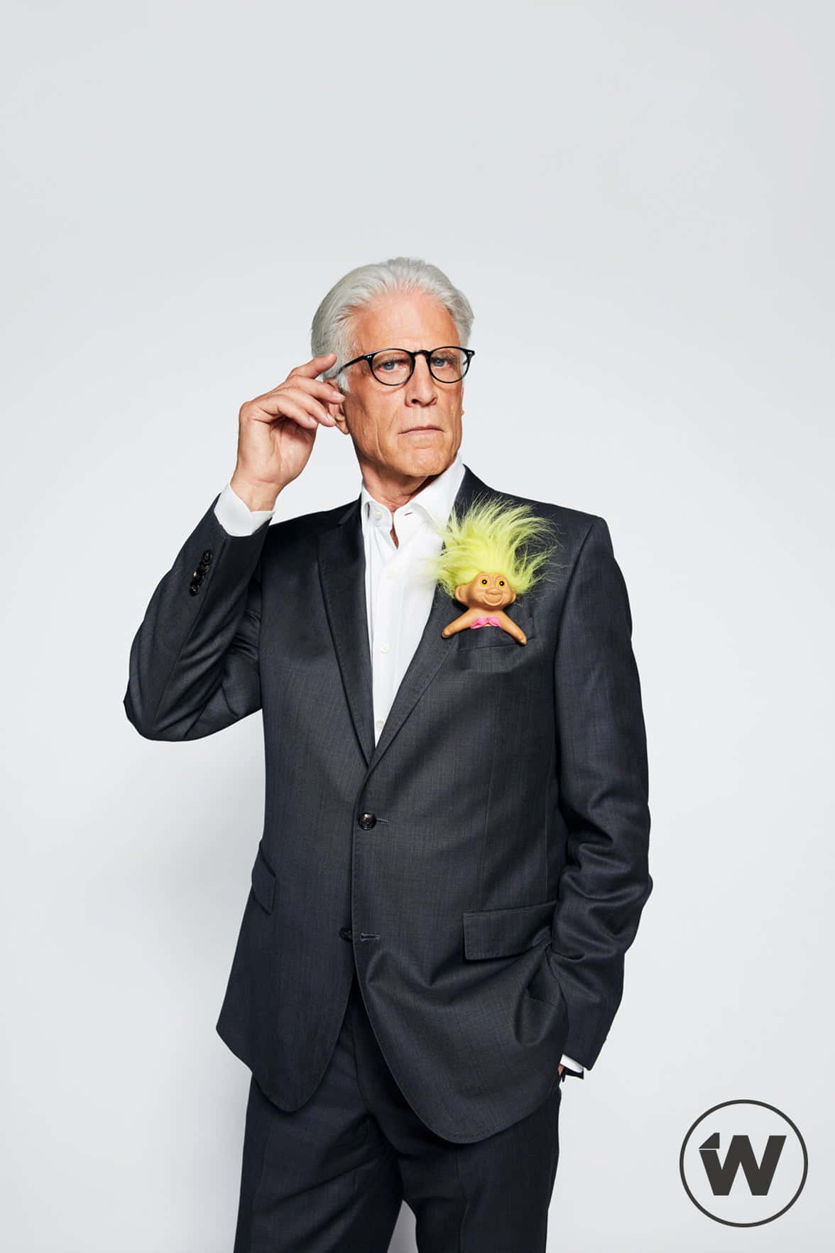 Ted Danson 桌布 壁紙 — 一名身穿深色西裝的男子正在調整眼鏡，他的翻領上放著一個亮黃色的巨魔娃娃，背景是純白色。