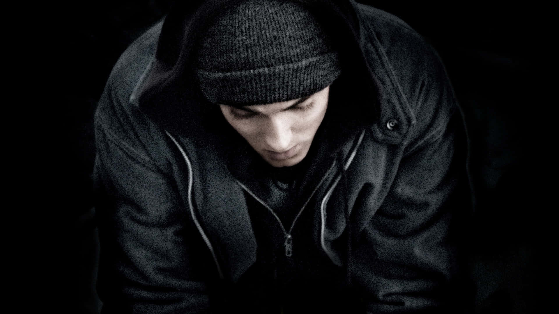 Eminem 桌布 壁紙 — 一名身穿深色連帽衫和冷帽的年輕男子在昏暗、陰鬱的環境中低頭。