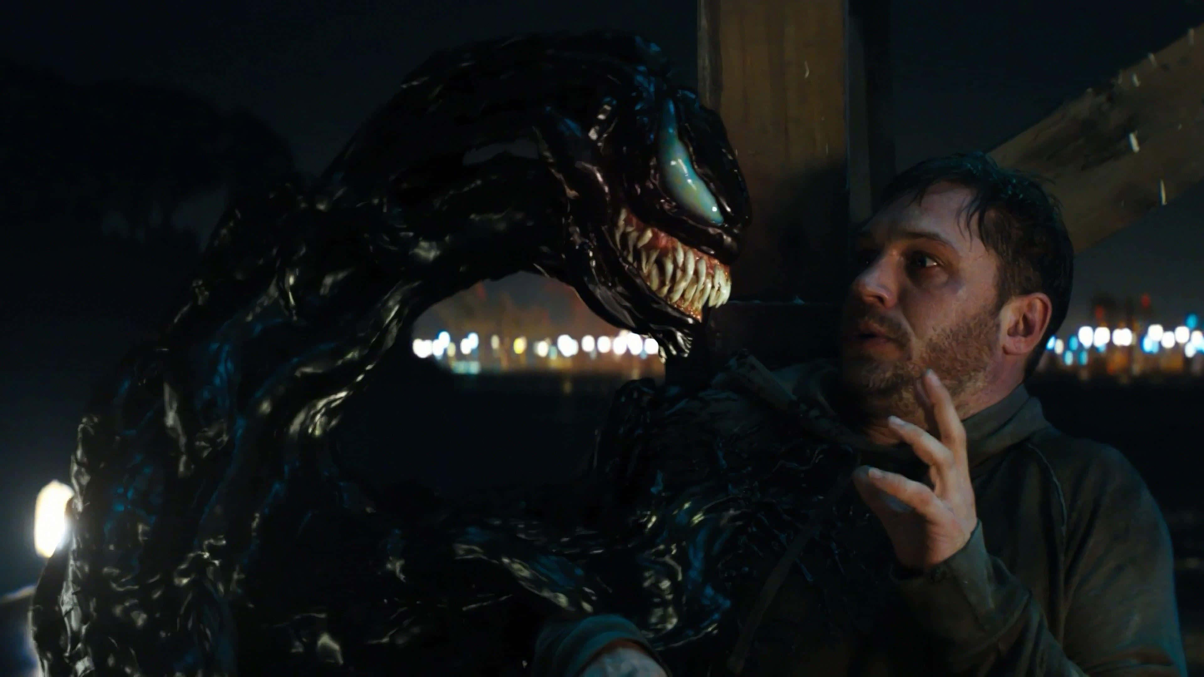 Tom Hardy Venom 桌布 壁紙 — 一名身穿深色連帽衫的男子在夜晚的城市燈光下，面對一個高聳的黑色共生體生物，它有著發光的眼睛和鋒利的牙齒。