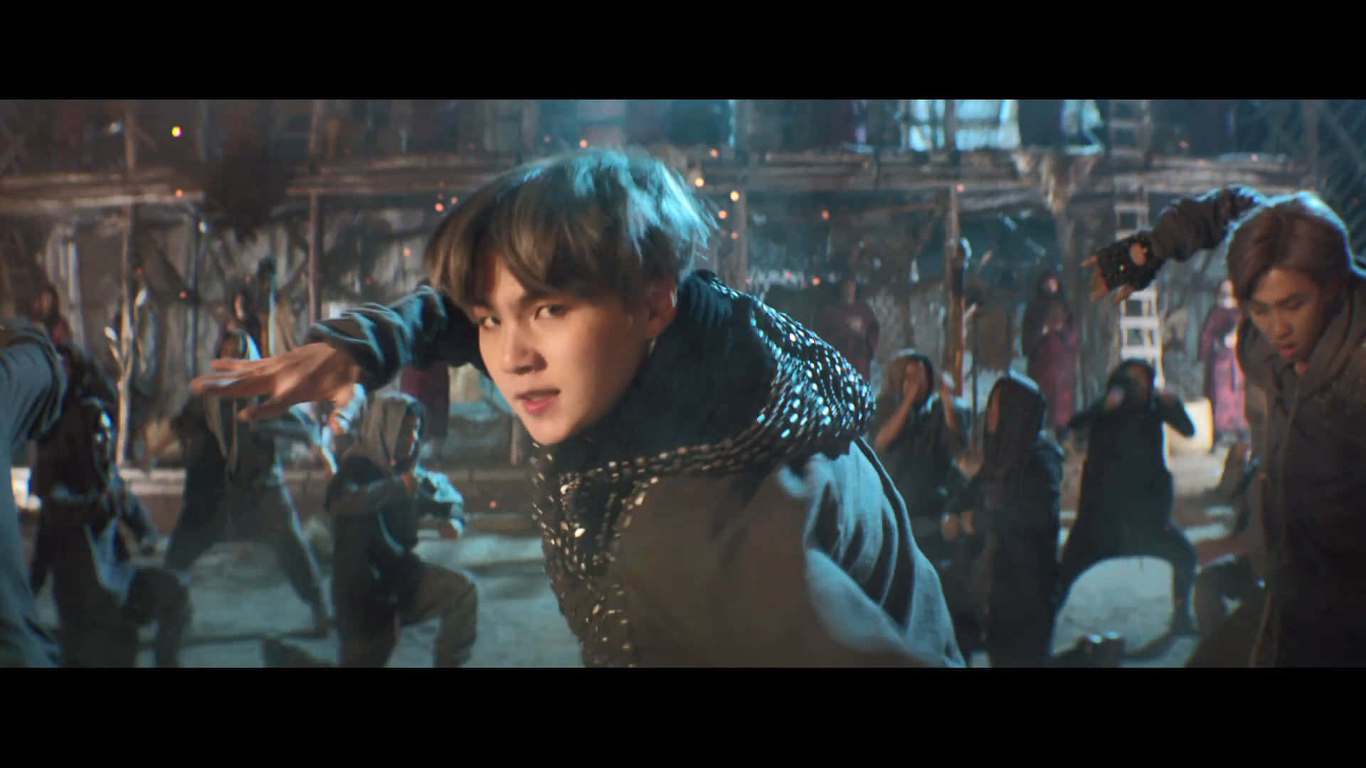 Bts Mv 桌布 壁紙 — 一名身穿深色鉚釘裝的年輕男子在昏暗、粗糙的城市環境中擺出動態姿勢，其他舞者也在移動。
