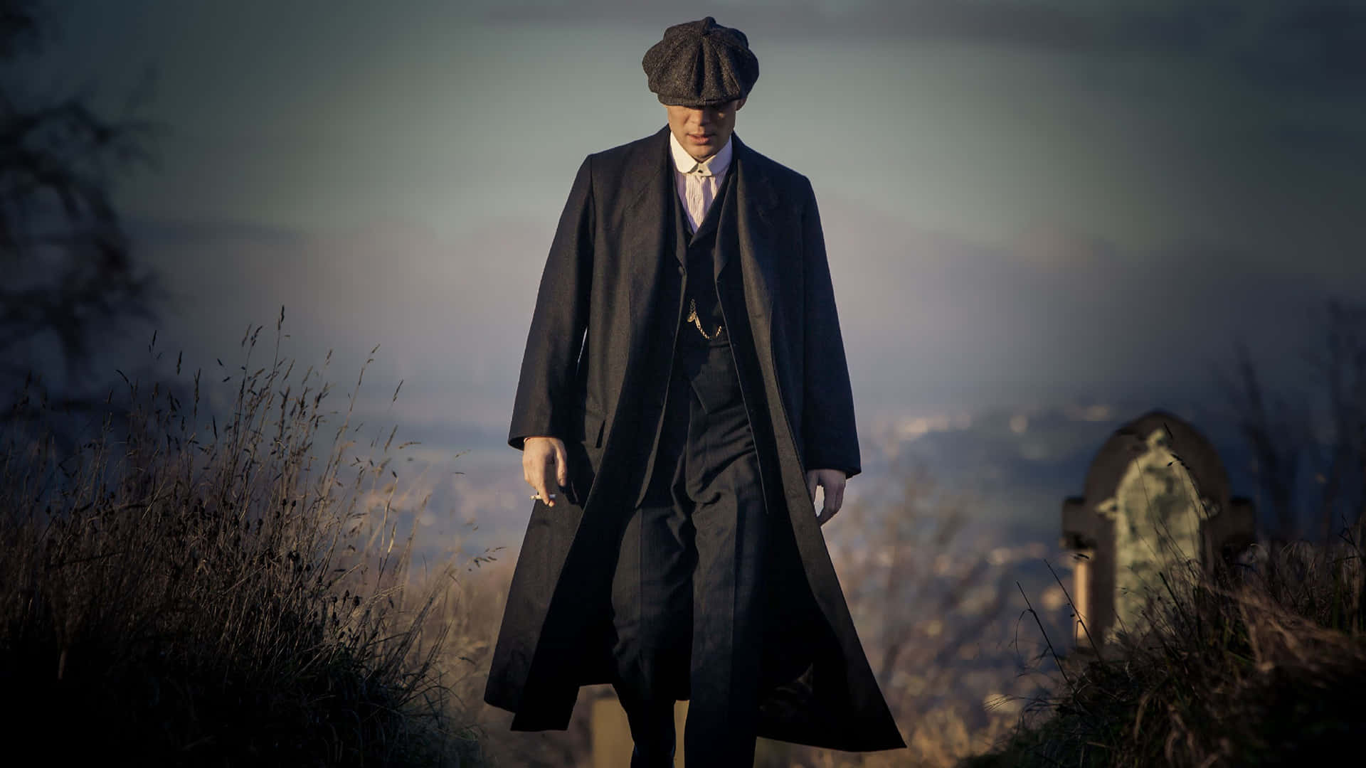 Peaky Blinders 桌布 壁紙 — 一名身穿深色風衣和扁帽的男子在黃昏時分莊嚴地走過霧氣瀰漫的墓地。
