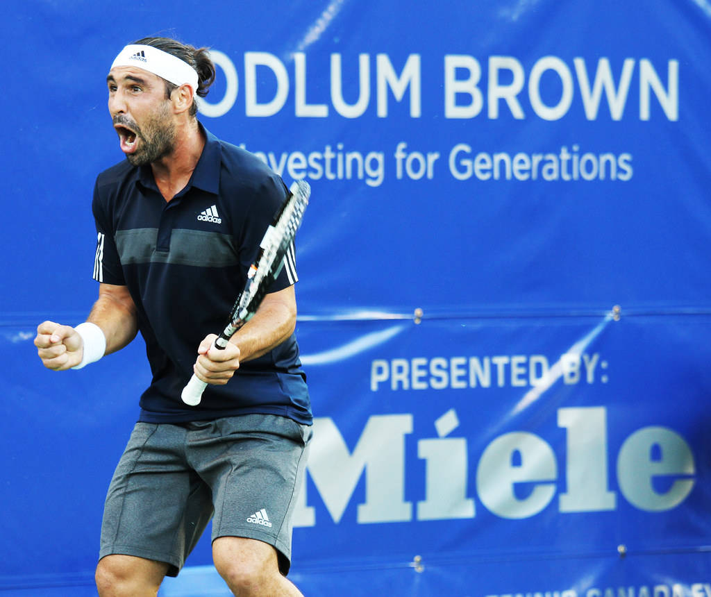 Marcos Baghdatis Cheering 壁紙 — 一名身穿深藍色Adidas球衣的男網球運動員，在鮮藍色的贊助商背景前，握緊拳頭、張開嘴巴，熱情地慶祝勝利。