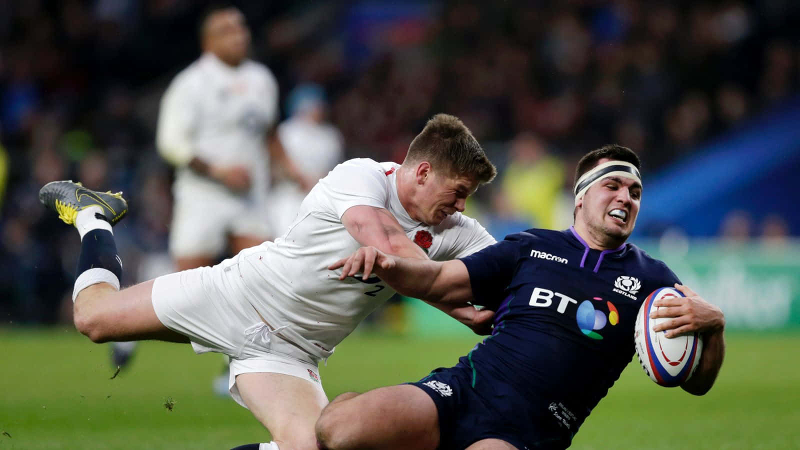 Scotland Rugby 桌布 壁紙 — 一名身穿深藍色球衣的欖球運動員在夜間比賽中緊緊抓住球，同時被一名身穿白色球衣的對手擒抱。