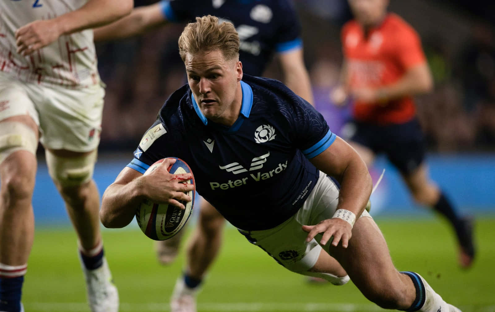 Scotland Rugby 桌布 壁紙 — 一名身穿深藍色球衣的欖球運動員在體育場燈光下進行激烈比賽時，抱著球向前飛撲。