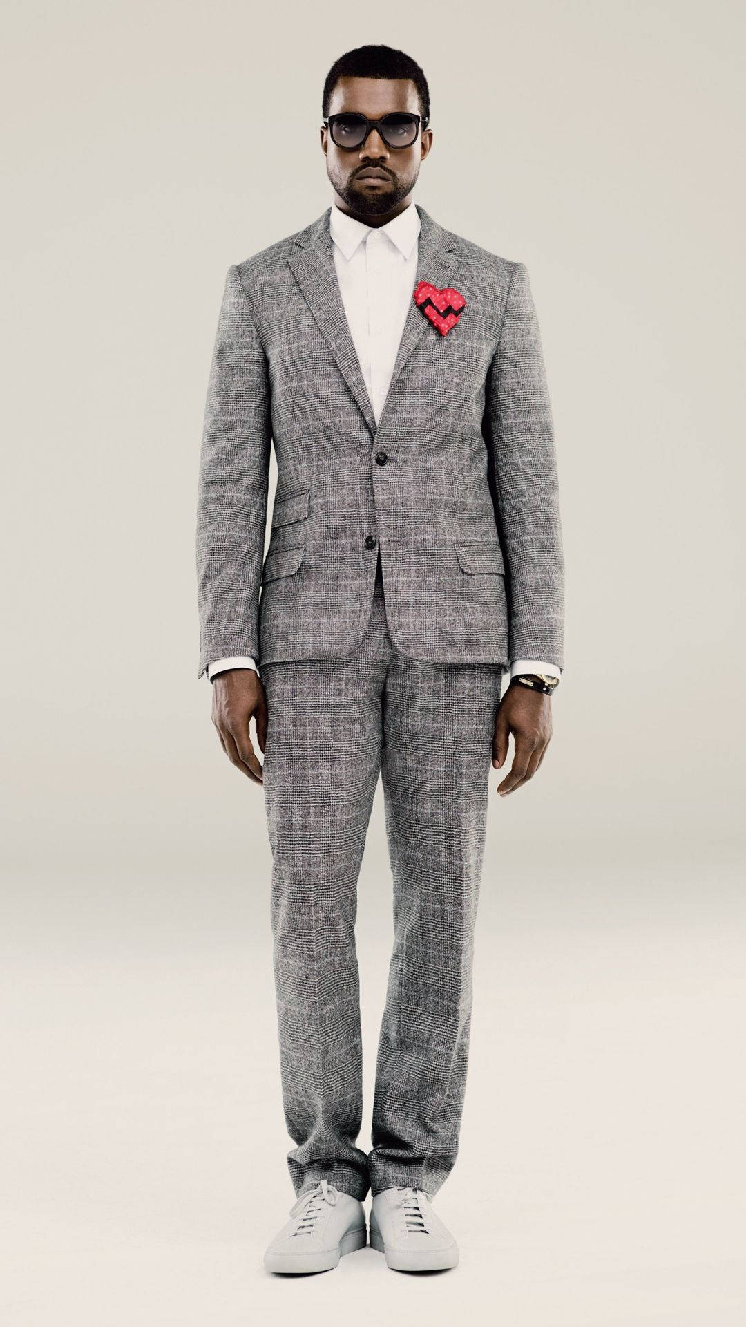 Kanye West In Grey Suit 壁紙 — 一名身穿灰色格紋西裝、佩戴紅色心形胸針的男子，自信地站在素色背景前。