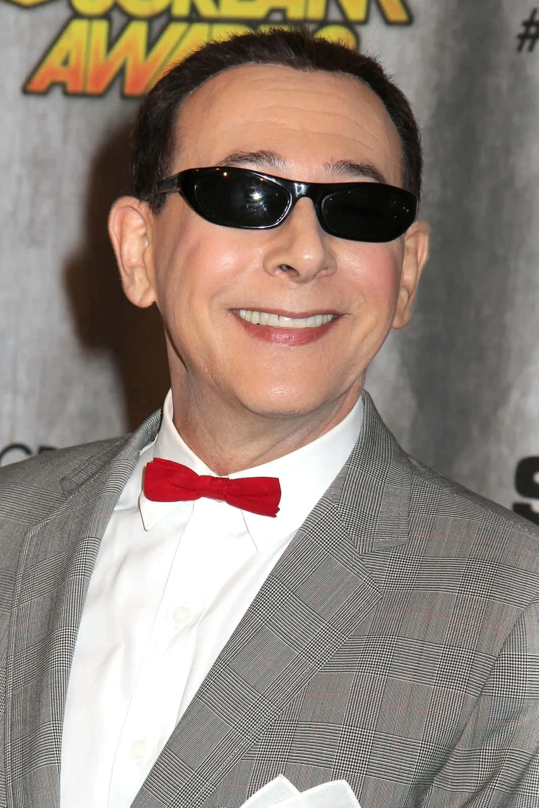 Paul Reubens 桌布 壁紙 — 一名身穿灰色格紋西裝和紅色領結的男子，在頒獎活動中戴著黑色太陽眼鏡微笑。