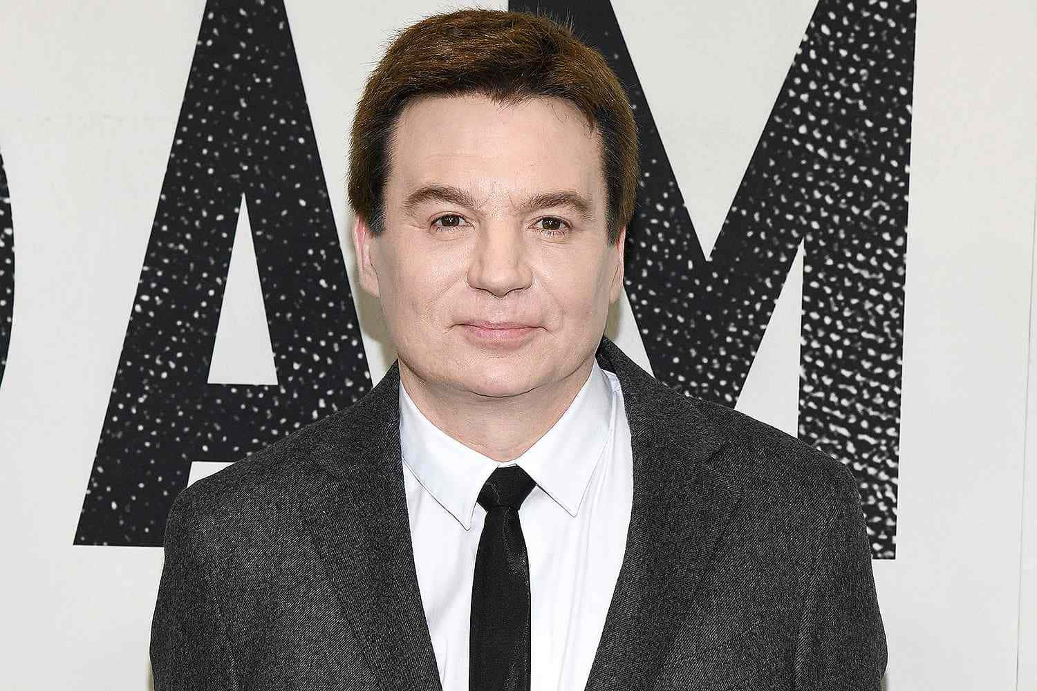Mike Myers 桌布 壁紙 — 一名身穿灰色西裝、打黑色領帶的男子站在大膽的黑白圖案背景前，背景上有大字母。