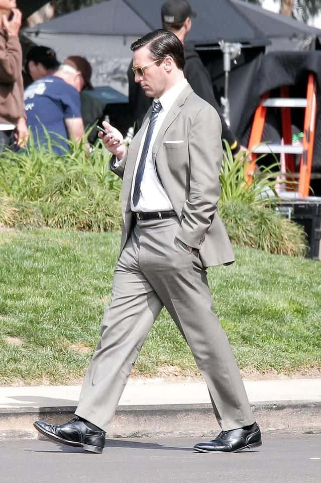 Jon Hamm 桌布 壁紙 — 一名身穿灰色西裝和太陽眼鏡的男子，專注地看著手機走在行人路上。