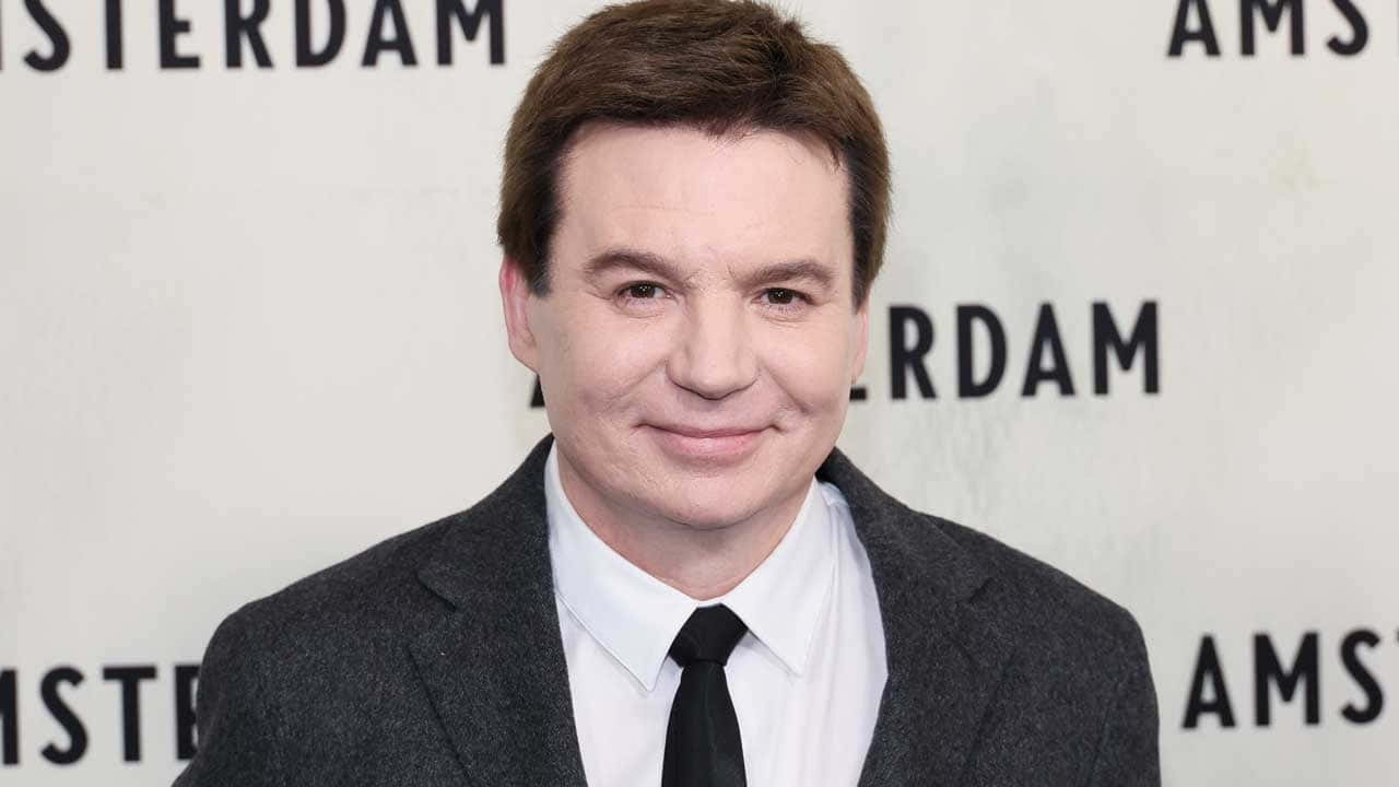Mike Myers 桌布 壁紙 — 一名身穿灰色西裝和黑色領帶的男子在阿姆斯特丹活動背景前對著鏡頭微笑。