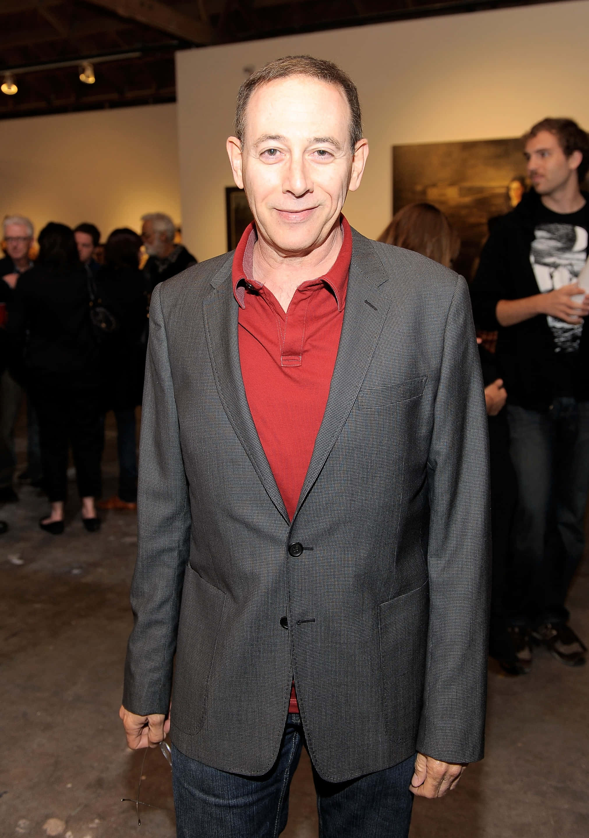 Paul Reubens 桌布 壁紙 — 一名身穿灰色西裝外套和紅色襯衫的男子在光線昏暗的畫廊中微笑著看向鏡頭，背景有藝術品和人群。