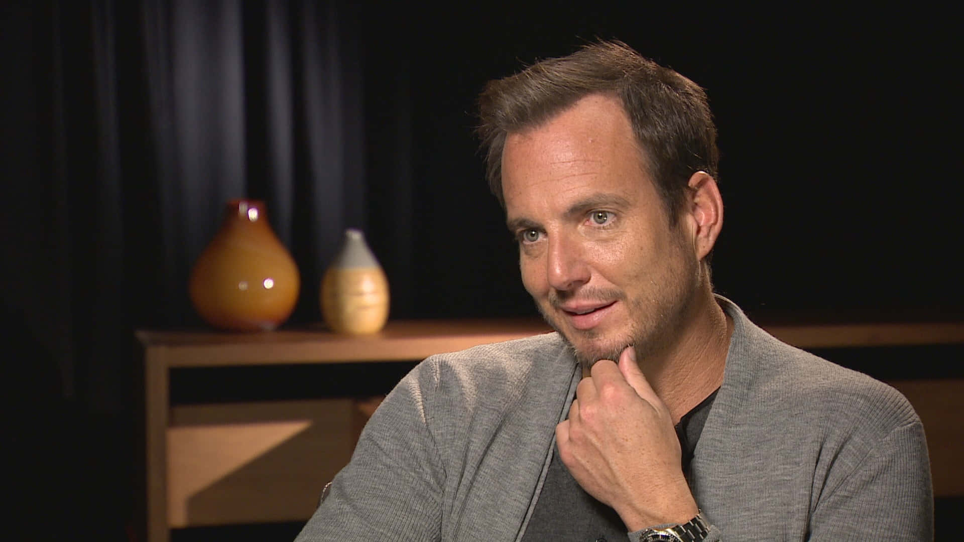 Will Arnett 桌布 壁紙 — 一名身穿灰色西裝外套的男子，在深色背景前，手托下巴沉思，旁邊有兩個琥珀色花瓶。