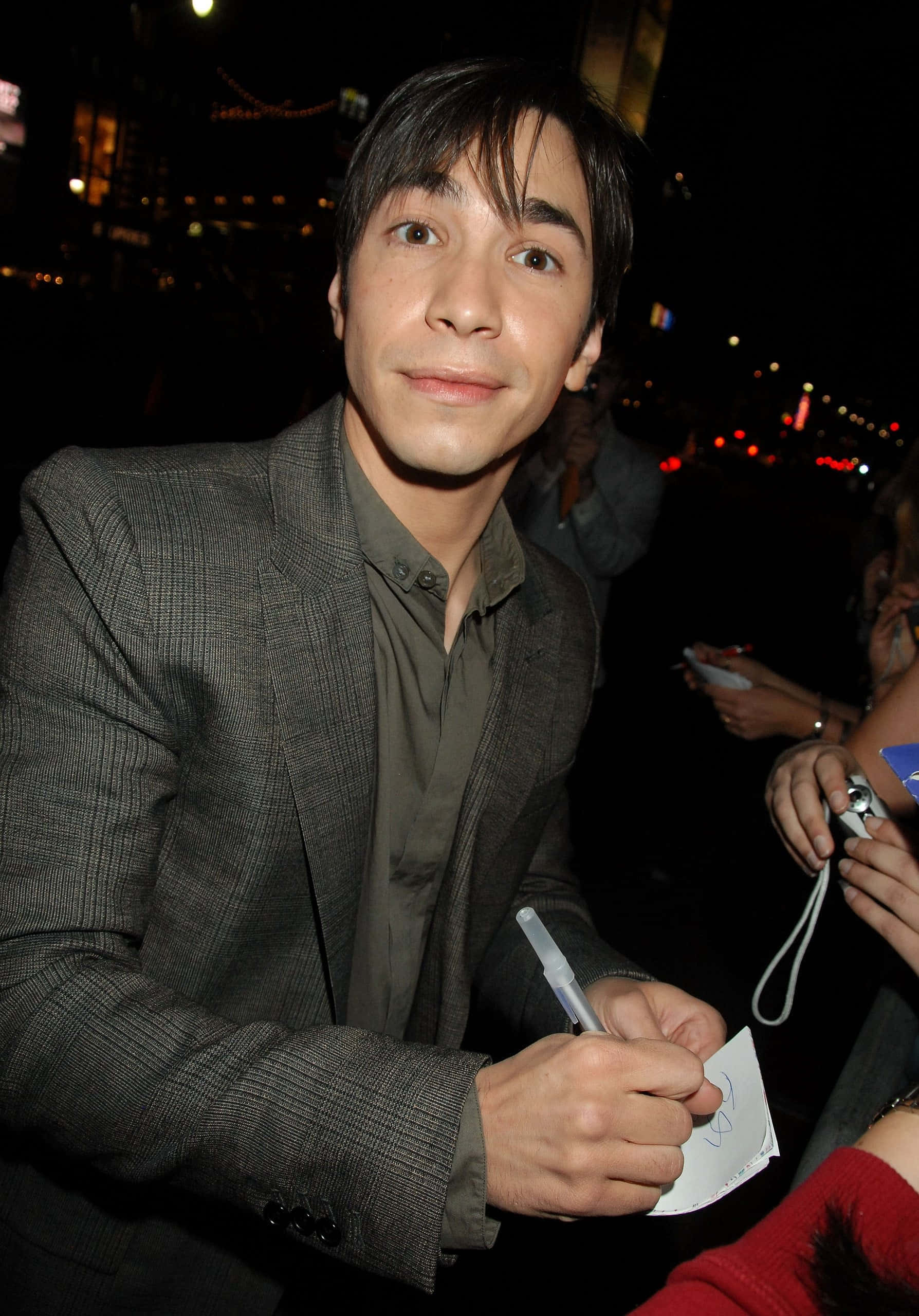 Justin Long 桌布 壁紙 — 一名身穿灰色西裝的男子在夜晚的城市燈光下微笑著為粉絲的物品簽名。