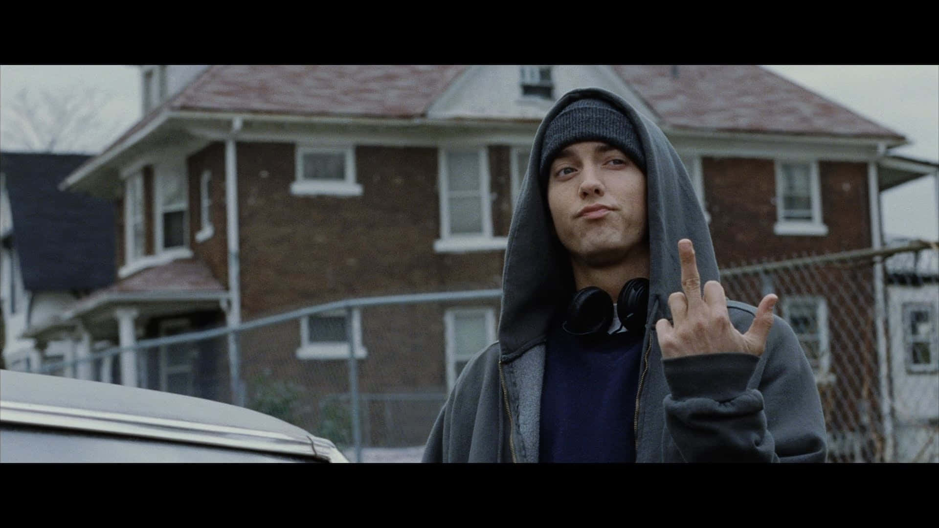 Eminem 桌布 壁紙 — 一名身穿灰色連帽衫和冷帽的年輕男子站在有鐵絲網圍欄的磚房前做手勢。