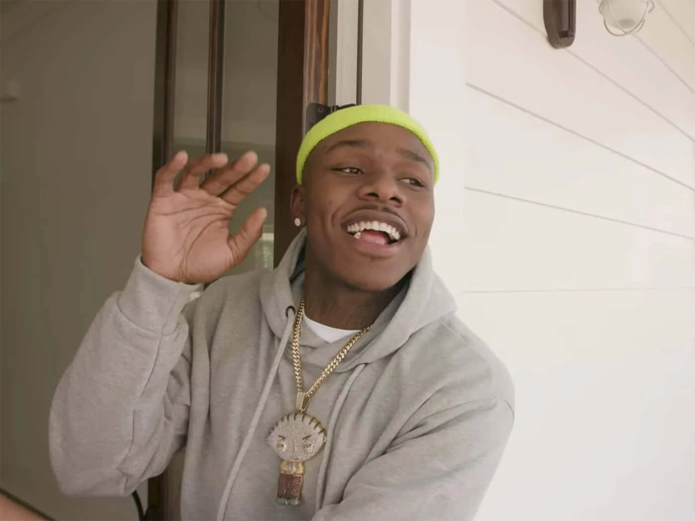 Dababy 桌布 壁紙 — 一名身穿灰色連帽衫和螢光黃色冷帽的年輕男子，戴著一條帶有臉部吊墜的大金鏈，愉快地揮手。