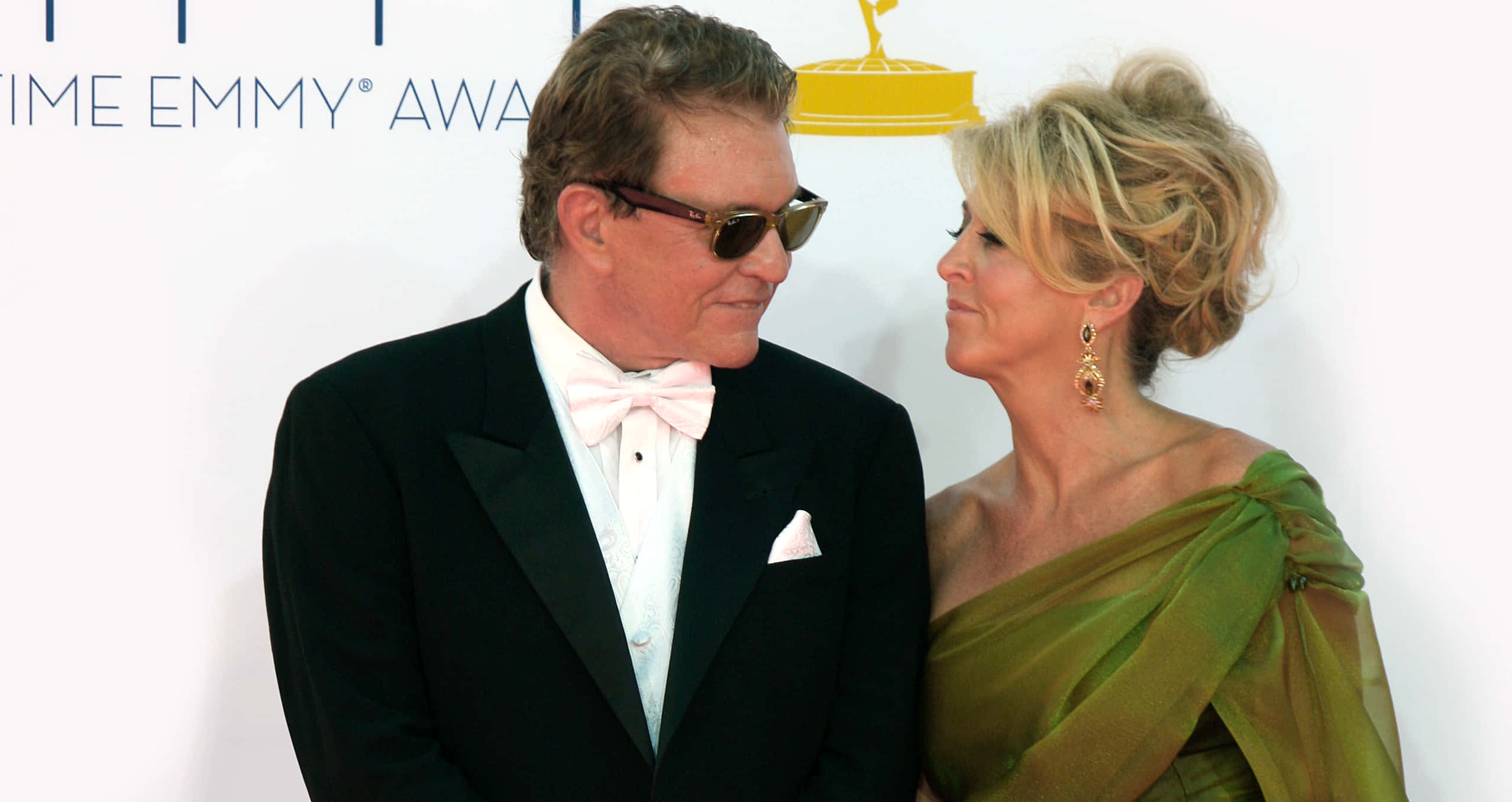 Tom Berenger 桌布 壁紙 — 一名身穿燕尾服、戴著太陽眼鏡和粉紅色領結的男子，在艾美獎頒獎典禮上站在一位身穿綠色禮服的女士旁邊。