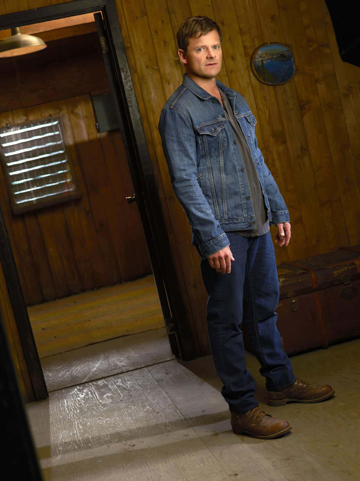 Steve Zahn 桌布 壁紙 — 一名身穿牛仔外套和牛仔褲的男子站在一個帶有復古行李箱和溫暖照明的鄉村風格木屋中。