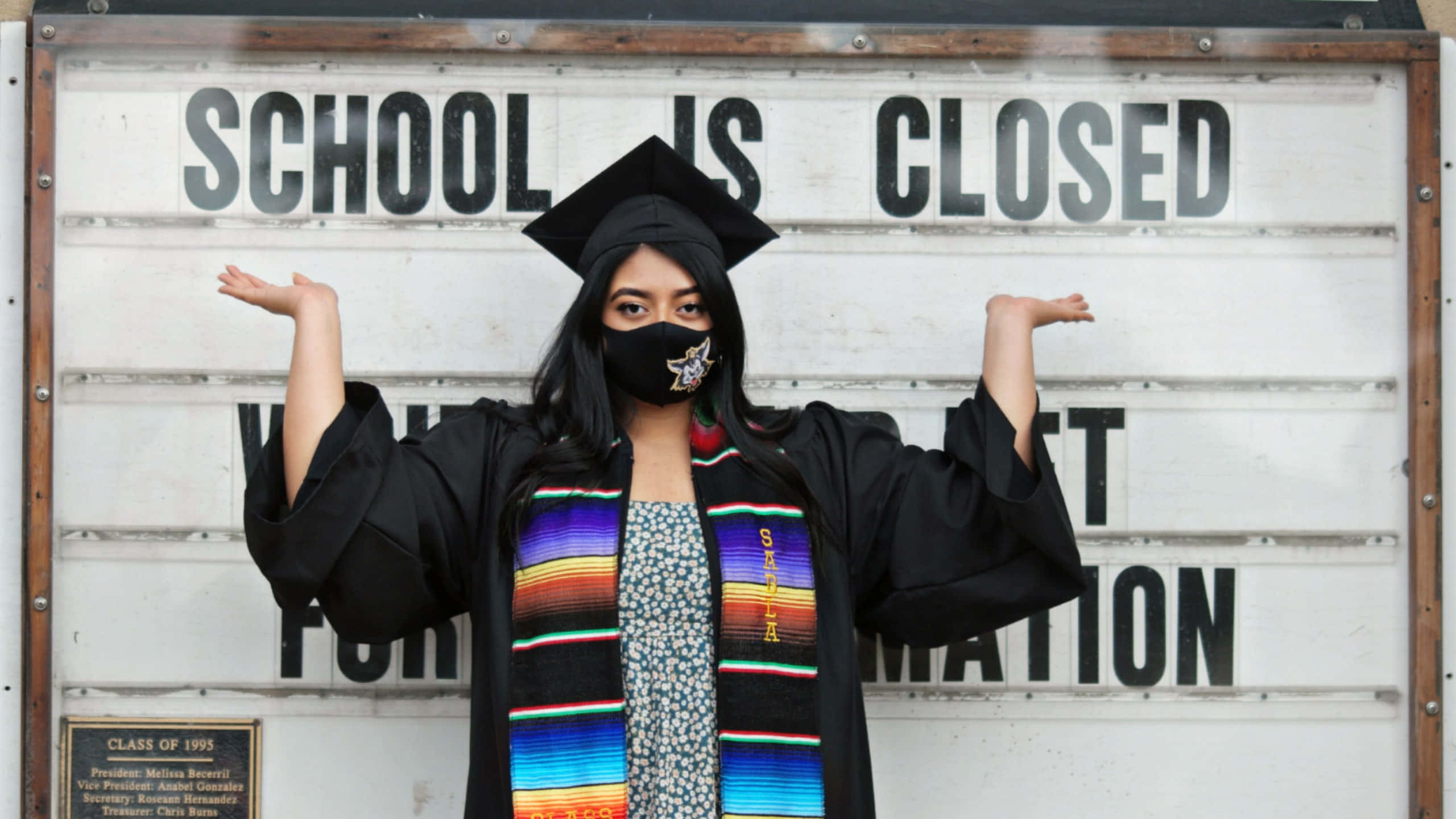 Graduation Ceremony 桌布 壁紙 — 一名身穿畢業袍、戴四方帽和彩色肩帶的畢業生，在「學校關閉」標誌前張開雙臂站立。