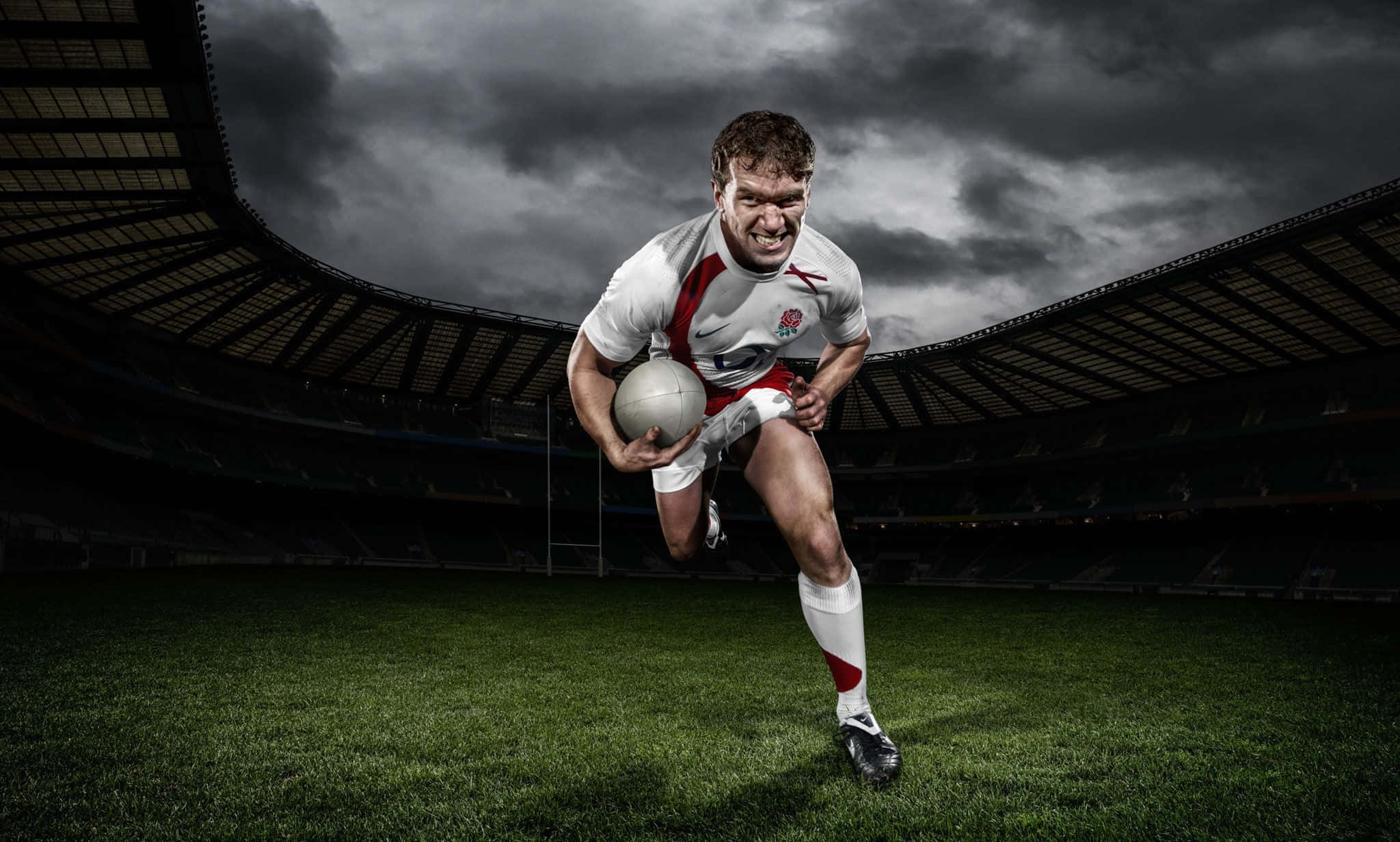 Rugby 桌布 壁紙 — 一名身穿白紅色球衣的欖球運動員在空無一人的球場上，在戲劇性的暴風雨天空下，持球衝過綠色草地。
