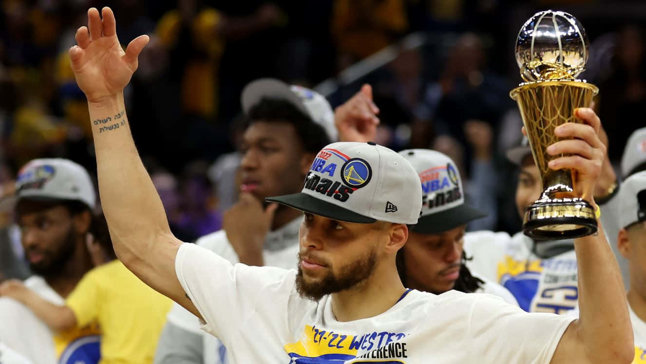 steph curry 桌布 壁紙 — 一名身穿白色T恤和帽子的籃球運動員高舉閃亮的NBA總決賽獎盃，向擠滿觀眾的場館內的球迷揮手。