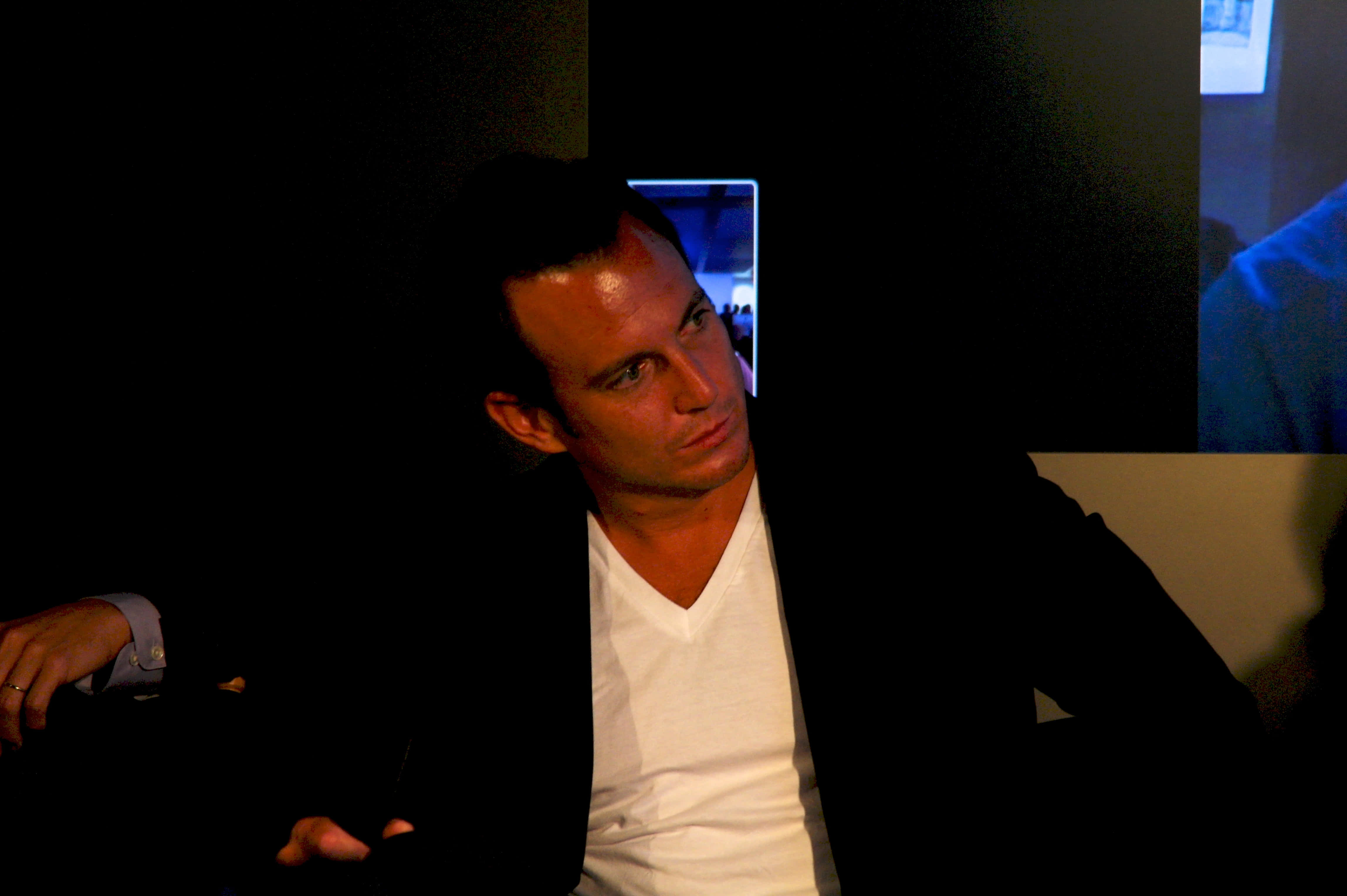 Will Arnett 桌布 壁紙 — 一名身穿白色V領和黑色西裝外套的男子坐在昏暗燈光下，目光專注，背後是黑暗陰影和發光的屏幕。