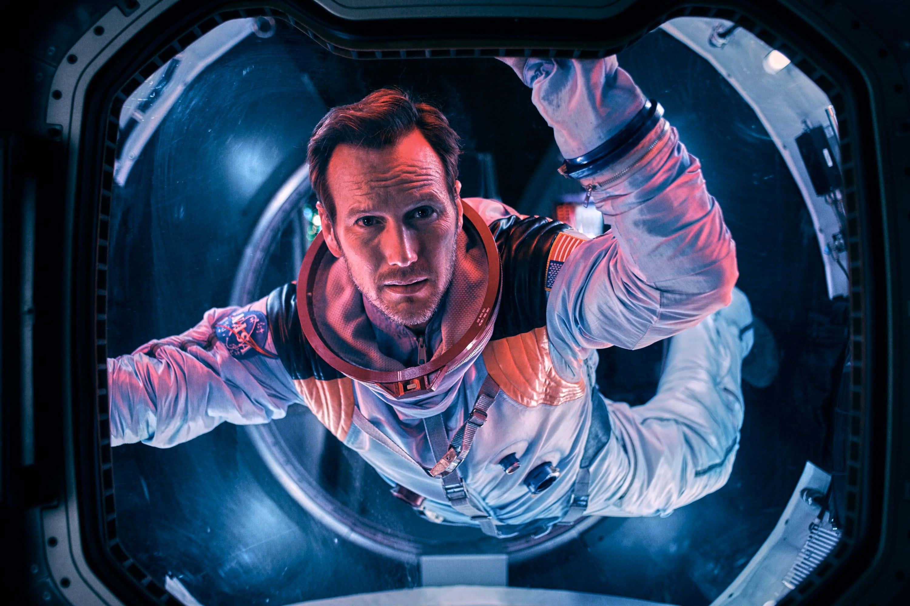 Patrick Wilson 桌布 壁紙 — 一名身穿白色和橙色太空衣的太空人，神情緊張地透過圓形太空船窗戶凝視著。