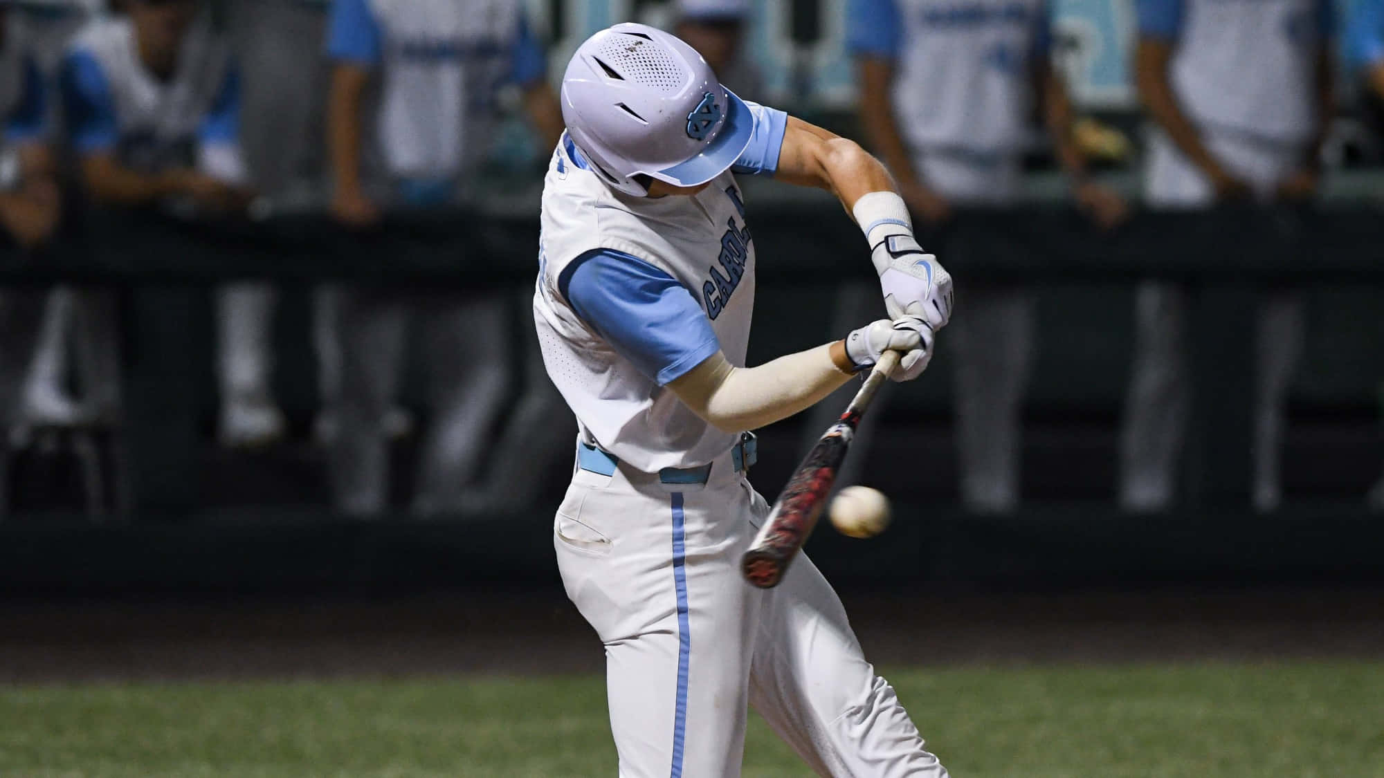 College Baseball 桌布 壁紙 — 一名身穿白色和藍色球衣的棒球手在球場燈光下揮棒擊球，背景中的隊友模糊不清。