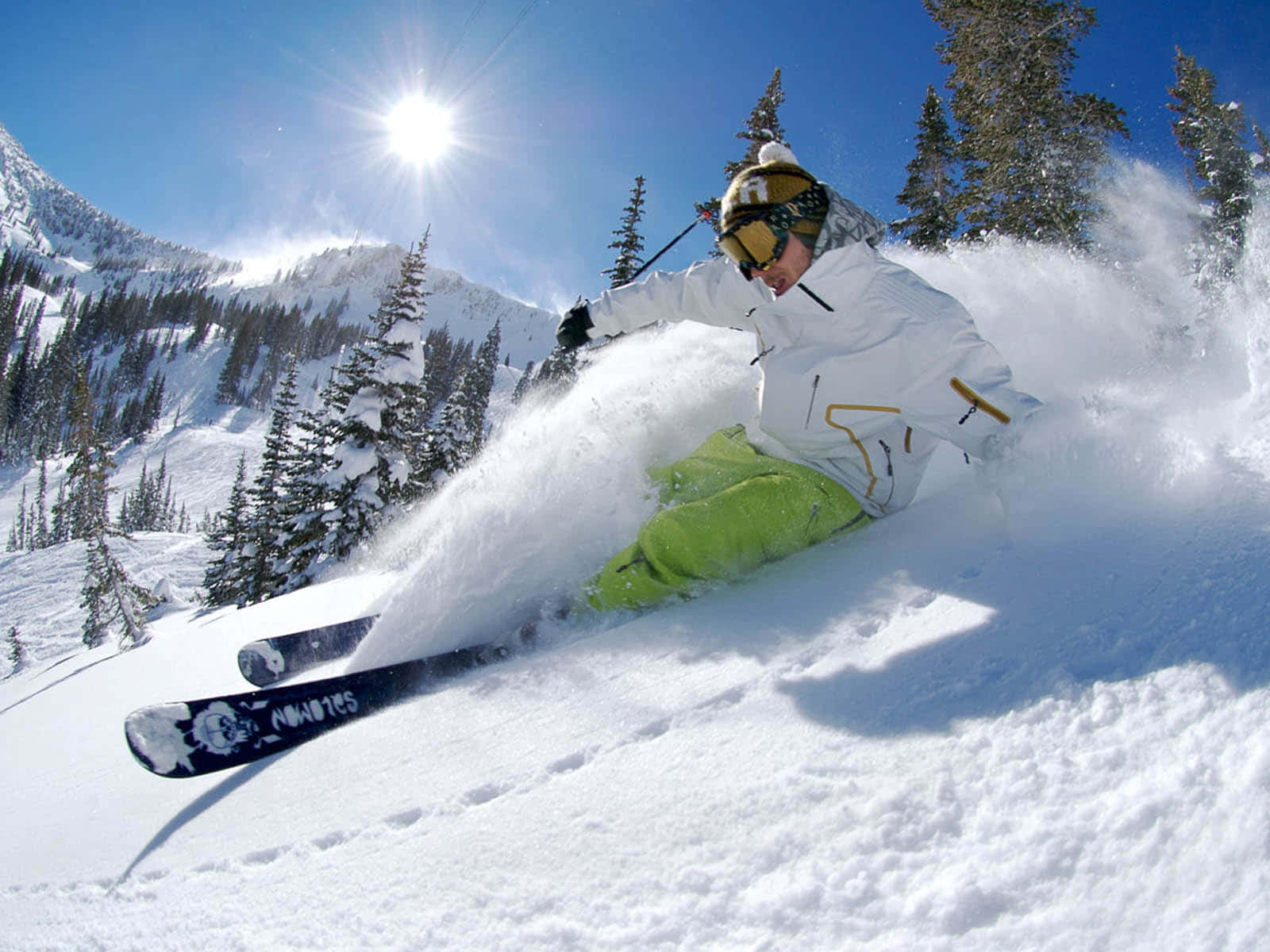Skiing 桌布 壁紙 — 一名身穿白色和青檸色裝備的滑雪者，在陽光明媚的雪坡上穿過新鮮的粉雪，背景是常青樹和山脈。