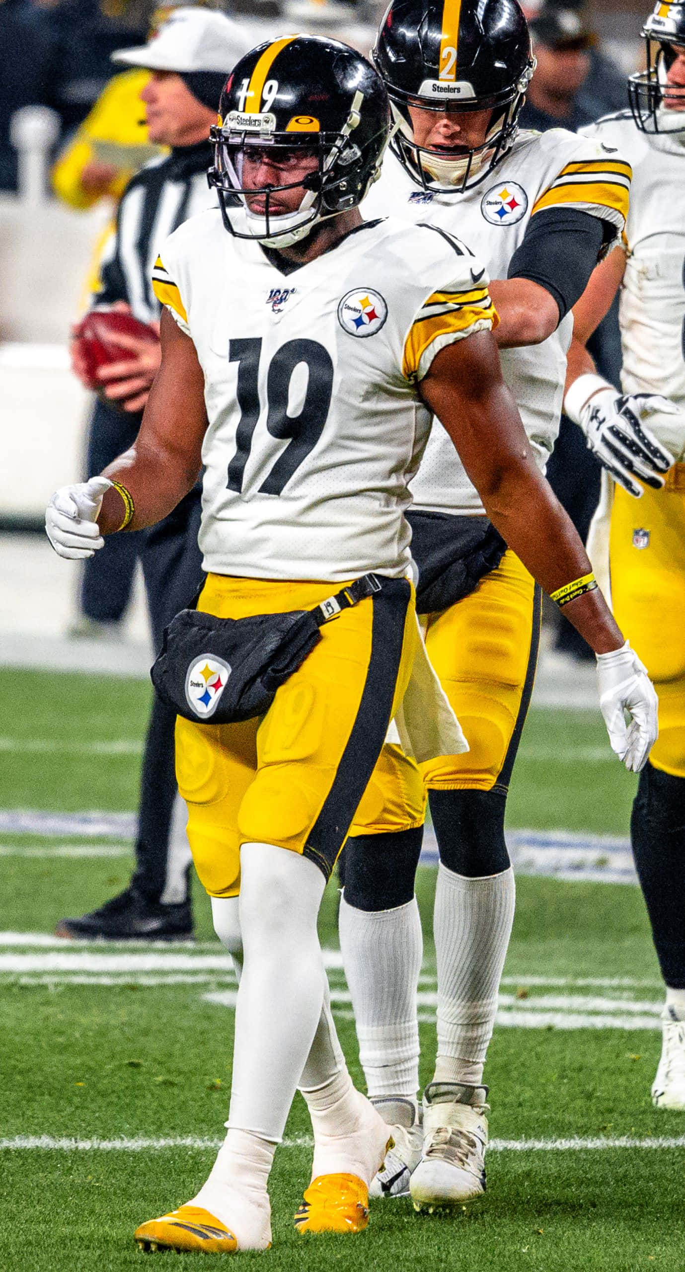 Juju Smith Schuster 桌布 壁紙 — 一名身穿白色和黃色球衣的匹茲堡鋼人隊美式足球員在比賽期間於綠色球場上行走，球場燈光照射。