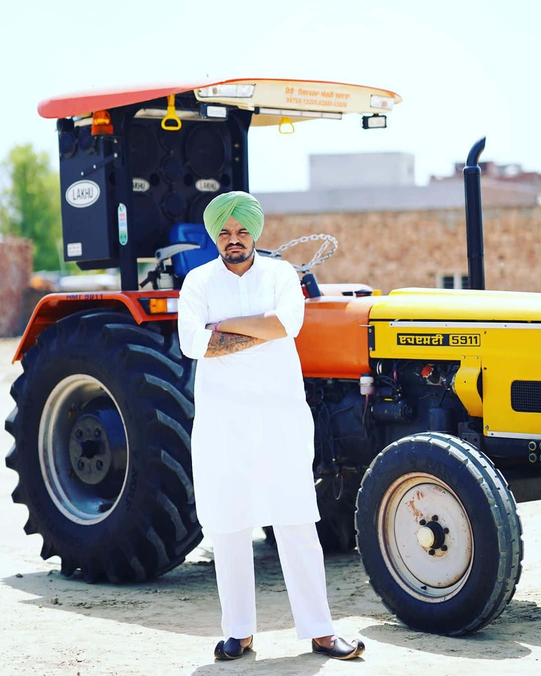 Sidhu Moose Wala 桌布 壁紙 — 一名身穿白色庫爾塔和綠色頭巾的男子，在明亮的日光下自信地交叉雙臂站在一輛黃色和橙色的拖拉機旁。