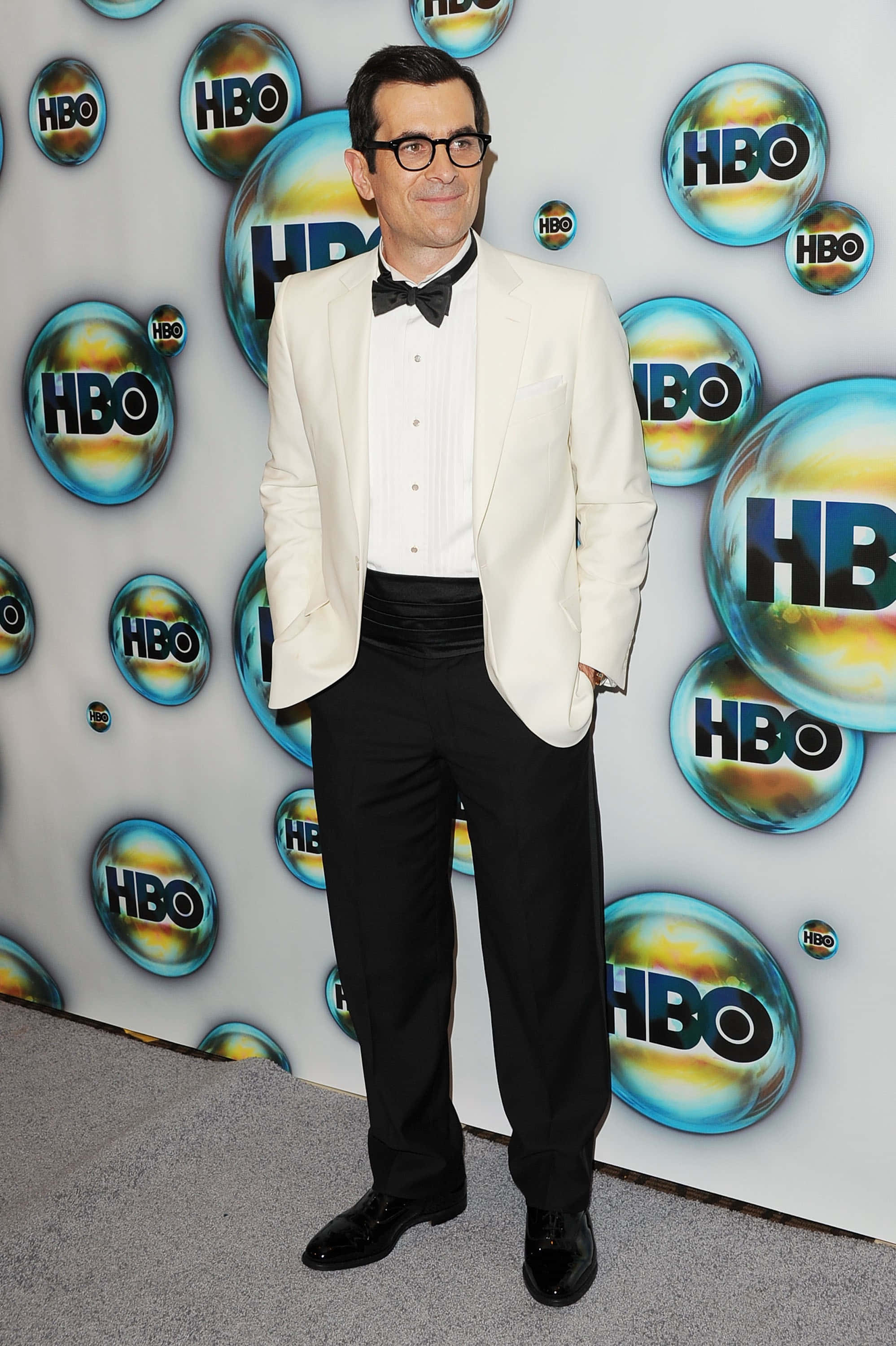 Ty Burrell 桌布 壁紙 — 一名身穿白色燕尾服外套和黑色領結的男子，自信地站在色彩繽紛的HBO標誌背景前。