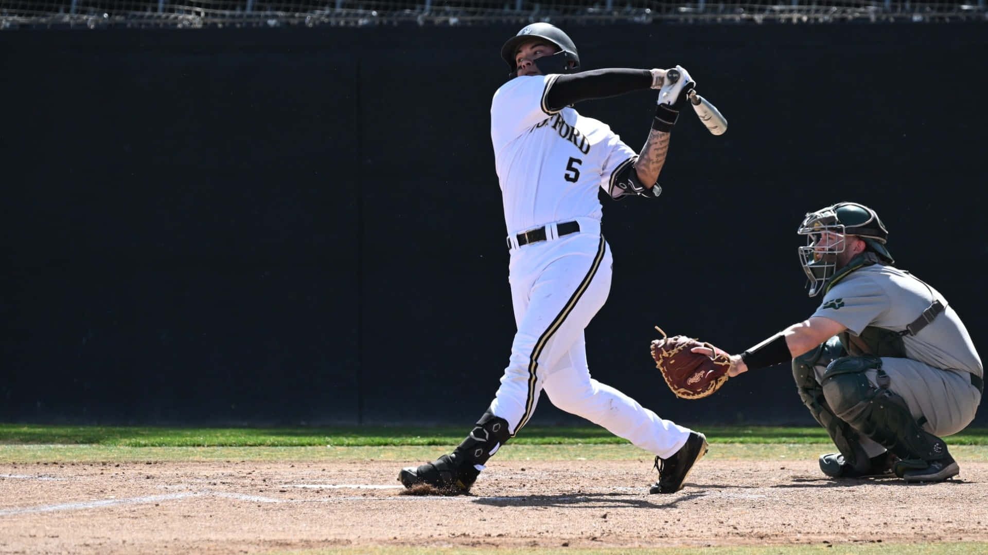 College Baseball 桌布 壁紙 — 一名身穿白色球衣的棒球手在本壘板揮棒，捕手全副裝備蹲在後面。