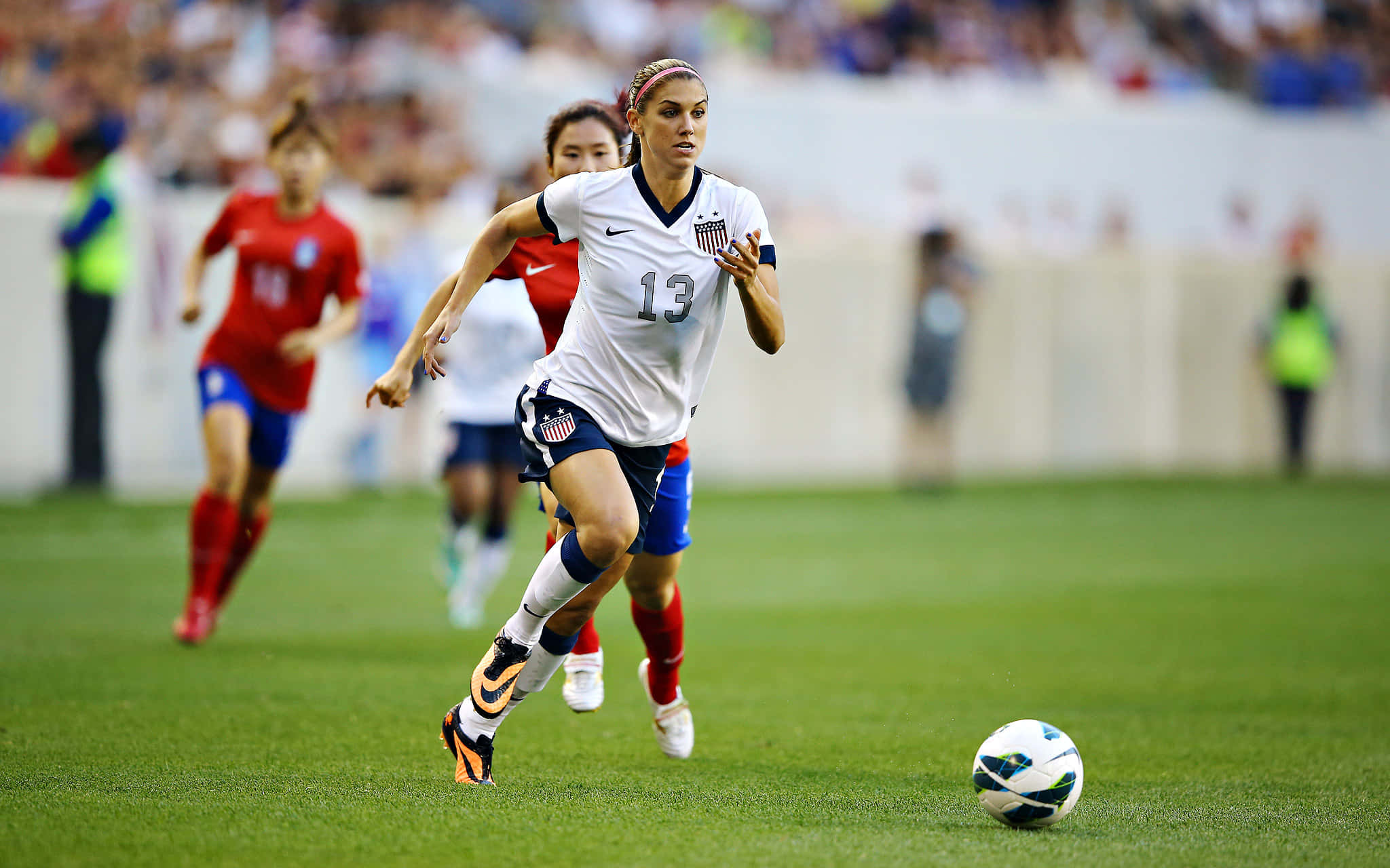 Alex Morgan 桌布 壁紙 — 一名身穿白色美國球衣的女足球員在充滿活力的綠色球場上專注地運球，對手在後面追趕。