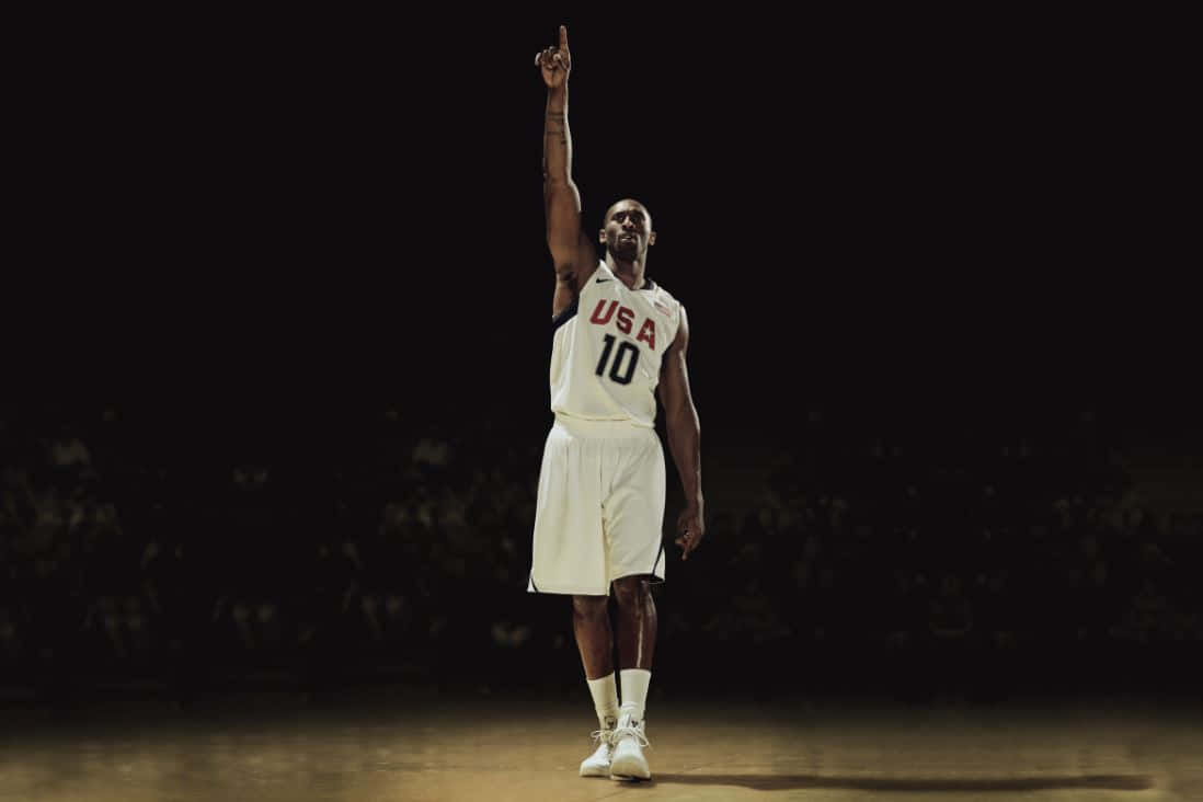 Kobe 桌布 壁紙 — 一名身穿白色美國隊10號球衣的籃球運動員獨自站在燈光昏暗的球場上，單手指向上方。