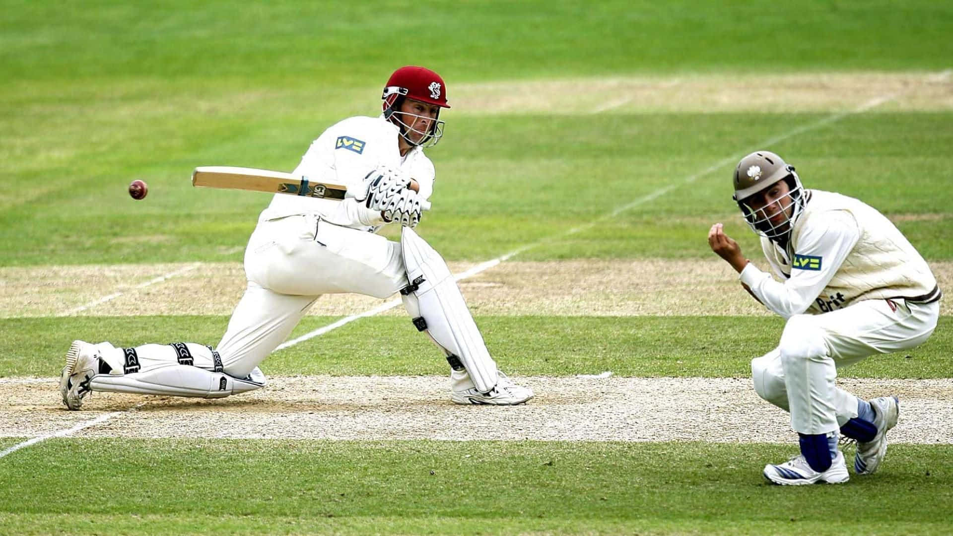Cricket 桌布 壁紙 — 一名身穿白色裝備的板球擊球手揮擊紅色球，而守門員在綠色場地上蹲伏準備。