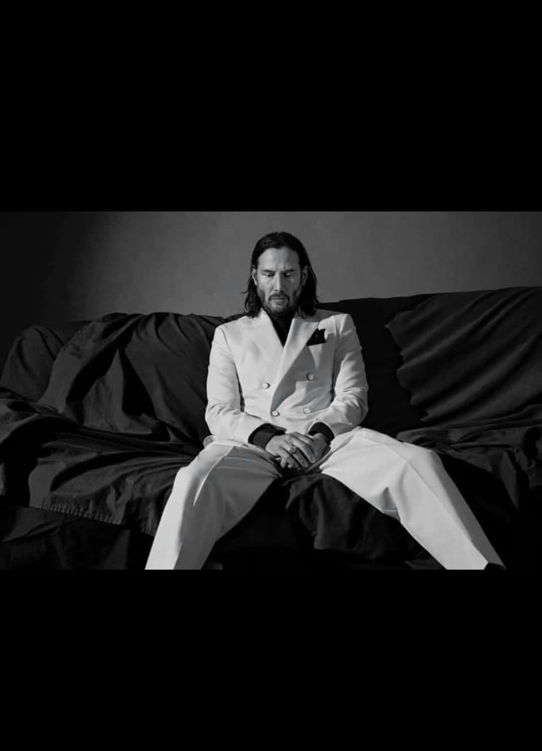 Keanu Reeves 桌布 壁紙 — 一名身穿白色西裝的男子莊嚴地坐在深色沙發上，呈現出鮮明的黑白工作室肖像。