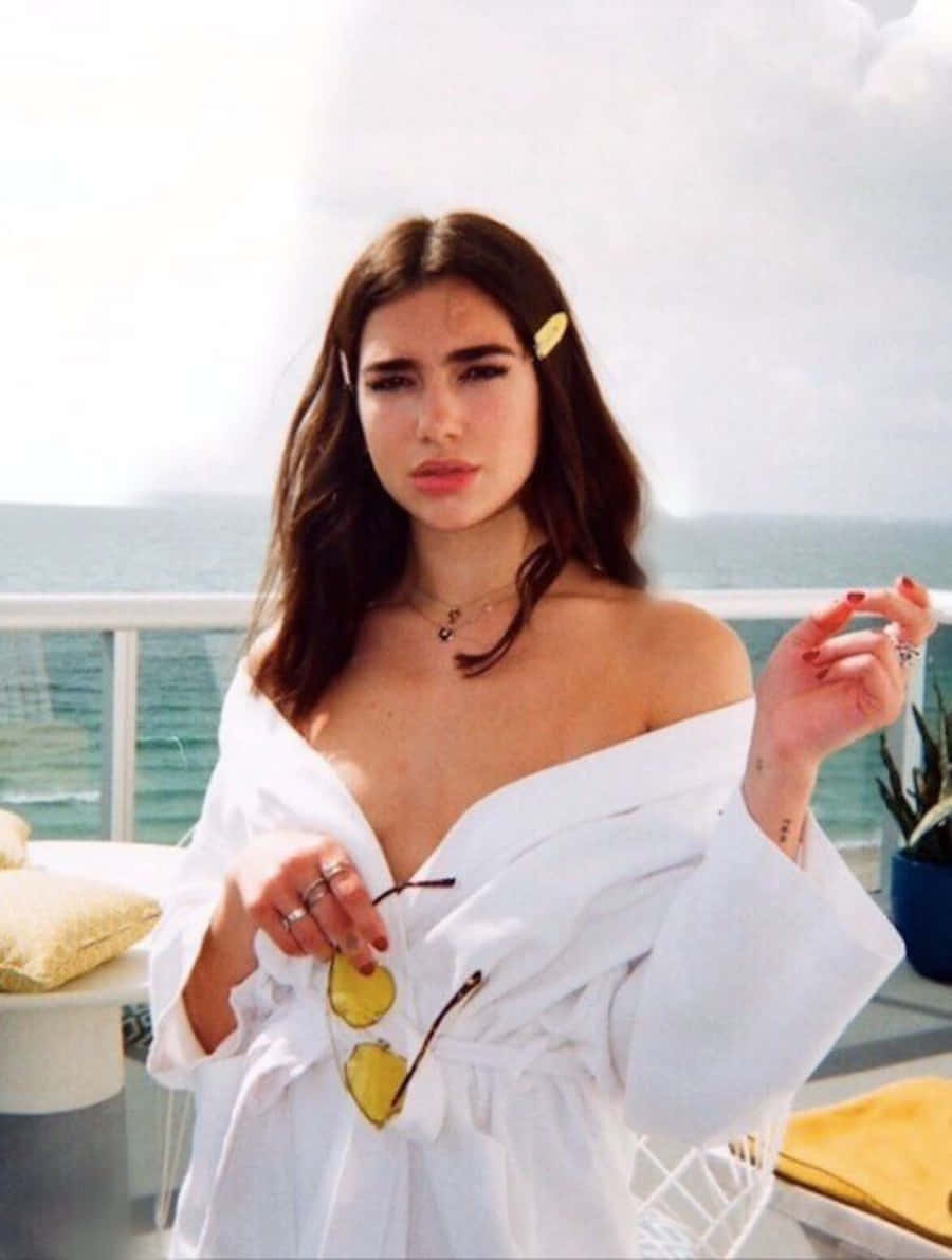 Dua Lipa 桌布 壁紙 — 一名身穿白色長袍的女子在海邊陽台上手持黃色太陽眼鏡，可欣賞海景。