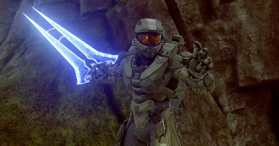 Halo Energy Sword 桌布 壁紙 — 一名身穿盔甲的斯巴達戰士在昏暗光線下的岩石地形中揮舞著發光的藍色能量劍。