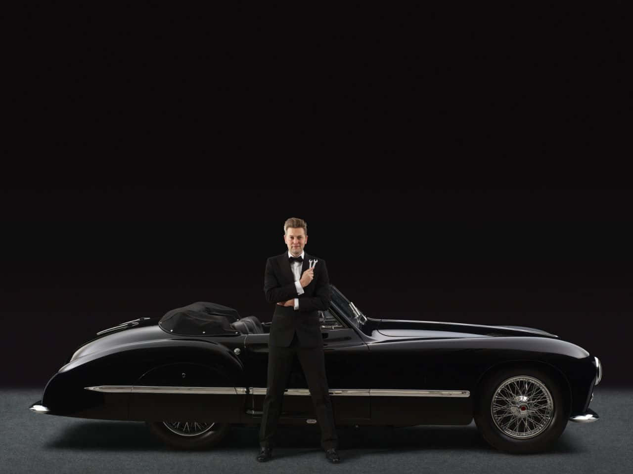 Black And White Vintage Car 桌布 壁紙 — 一名身穿禮服的男士自信地站在一輛時尚的黑色復古敞篷車旁，背景是深色攝影棚。