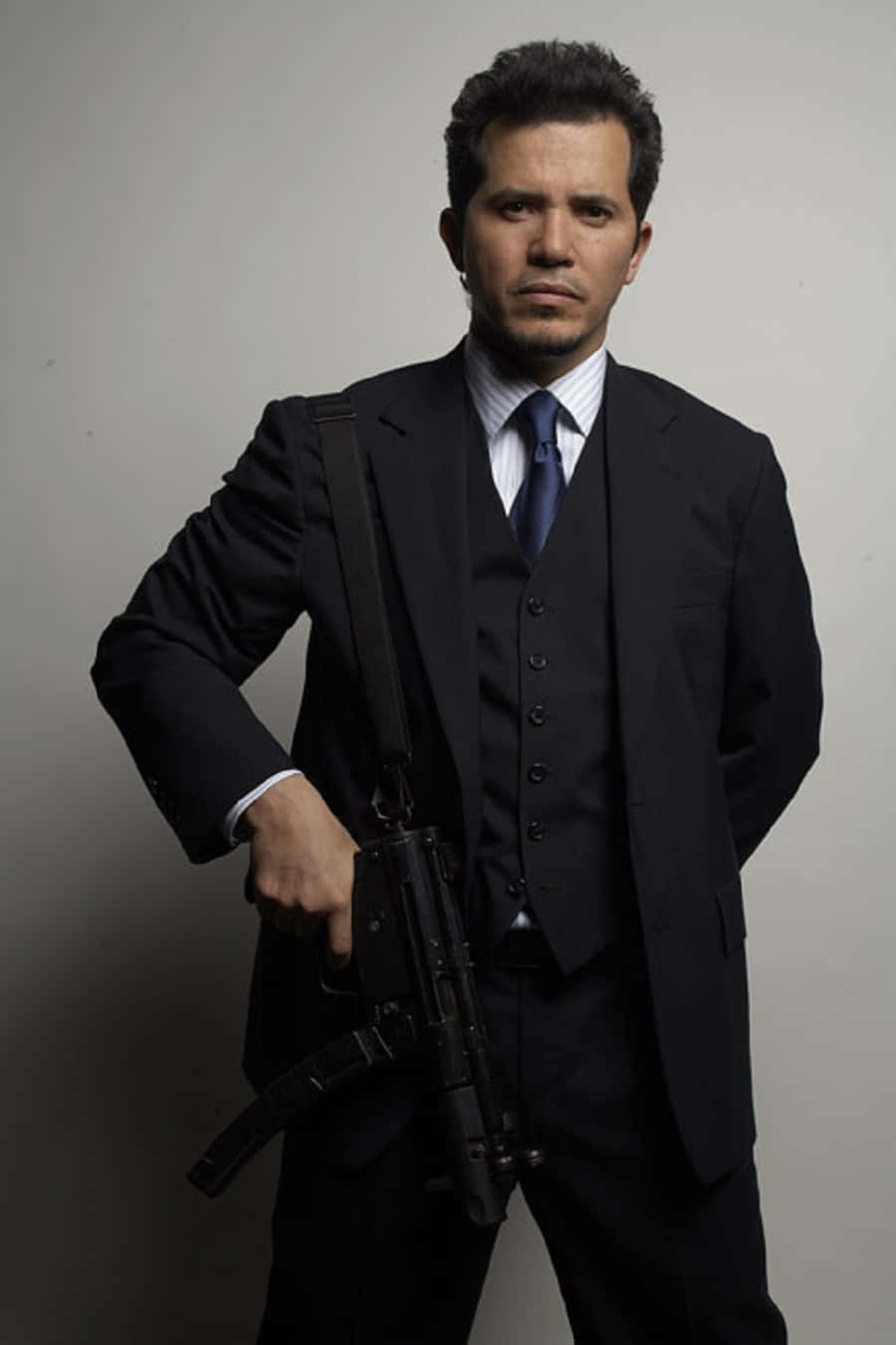 John Leguizamo 桌布 壁紙 — 一名身穿筆挺黑色西裝的男子，在純灰色背景前手持突擊步槍，表情嚴肅。