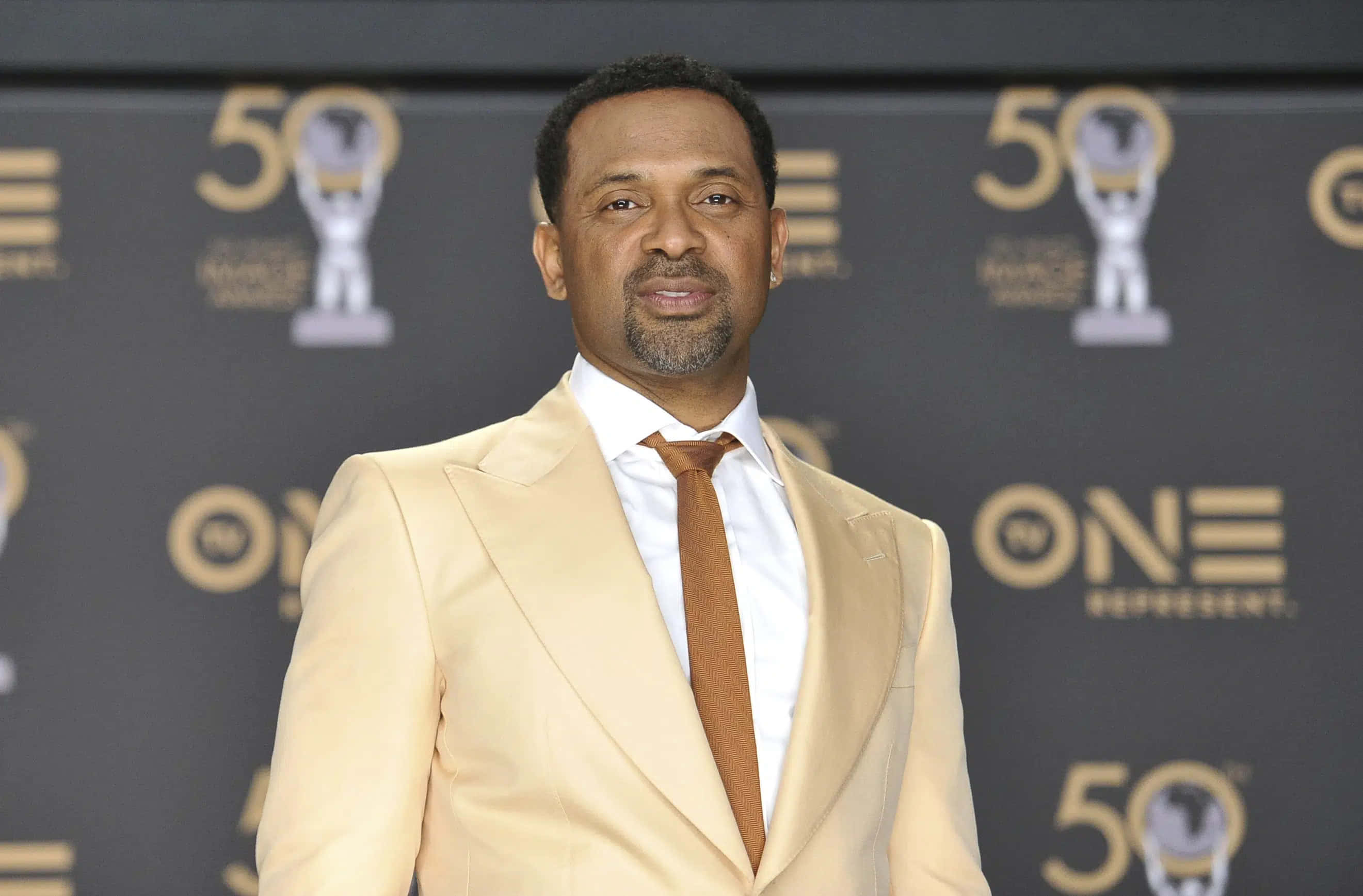 Mike Epps 桌布 壁紙 — 一名身穿米色西裝和棕色領帶的男子自信地站在黑色背景前，背景上有金色的「50」和獎盃標誌。