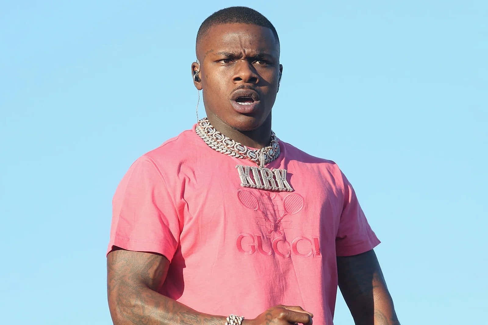 Dababy 桌布 壁紙 — 一名身穿粉紅色Gucci T恤、印有「KIRK」及網球拍標誌的男子，在清澈的藍天下戴著一條粗鏈條項鍊。