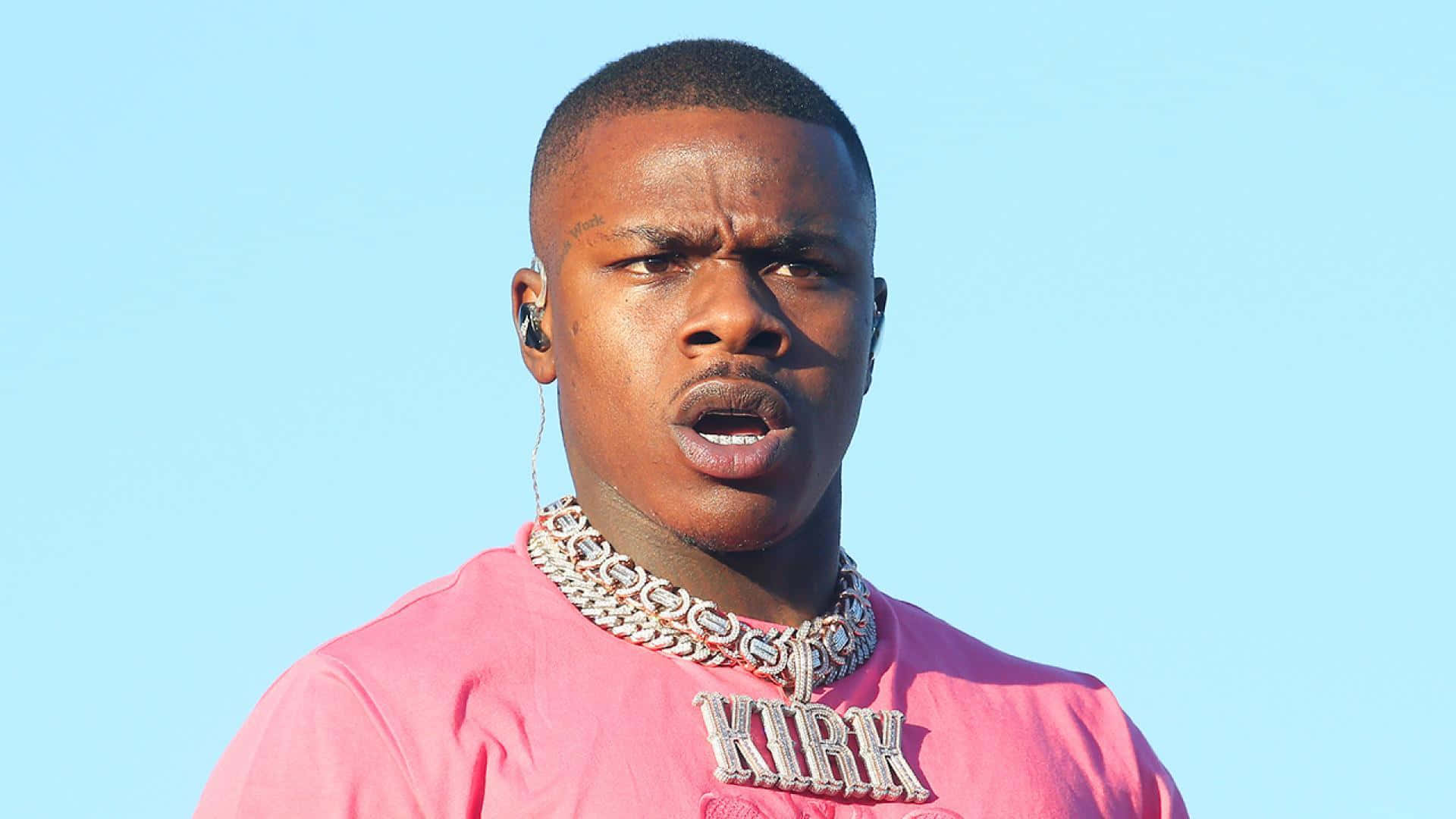 Dababy 桌布 壁紙 — 一名身穿粉紅色襯衫和粗鏈條項鍊的男子在清澈的藍天下專注地看著鏡頭。