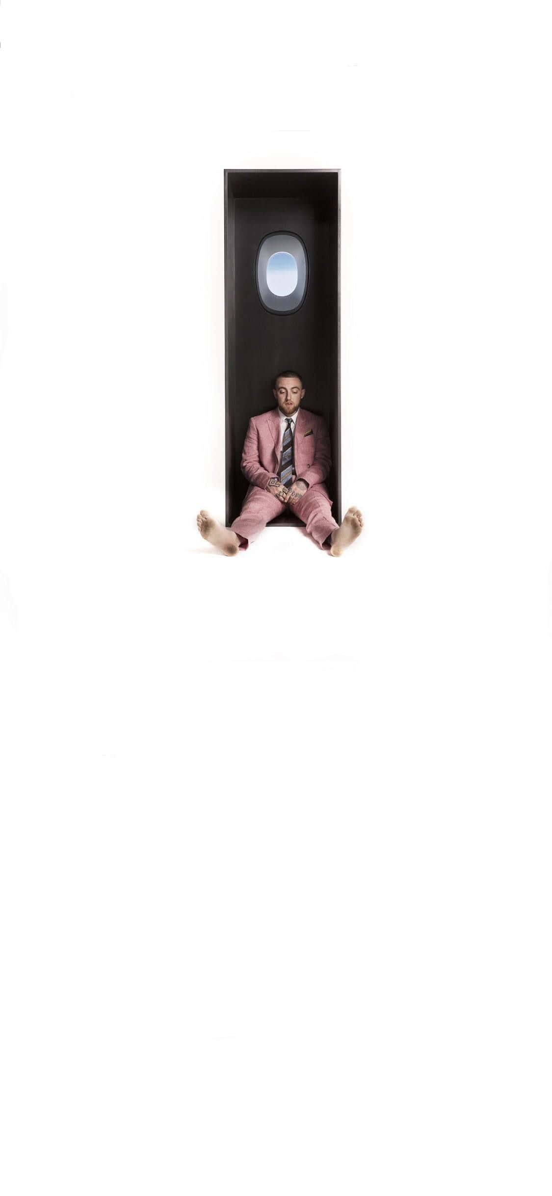 Mac Miller 桌布 壁紙 — 一名身穿粉紅色西裝的男子赤腳坐在一個狹窄黑暗的隔間內，隔間有一個小圓窗，可以看到藍天。
